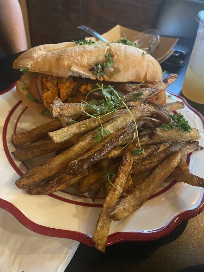 A plate of crispy French fries topped with fresh herbs and a sandwich with bread, fried chicken, and vegetables, served with a drink in the background.