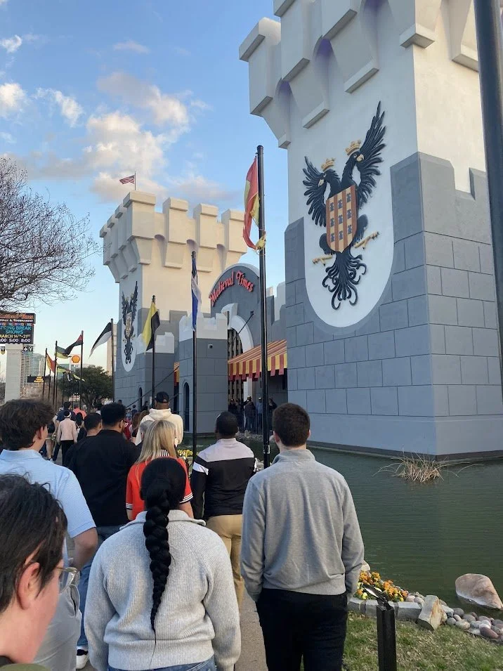 People waiting in line outside a castle-themed attraction with banners, flags, and a large shield emblem on the wall.
