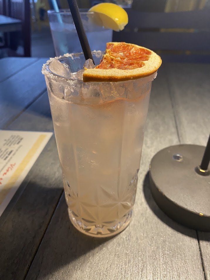 A tall glass of pale pink cocktail garnished with a lemon wedge and a slice of grapefruit, served with a black straw on a wooden table at an outdoor seating area.