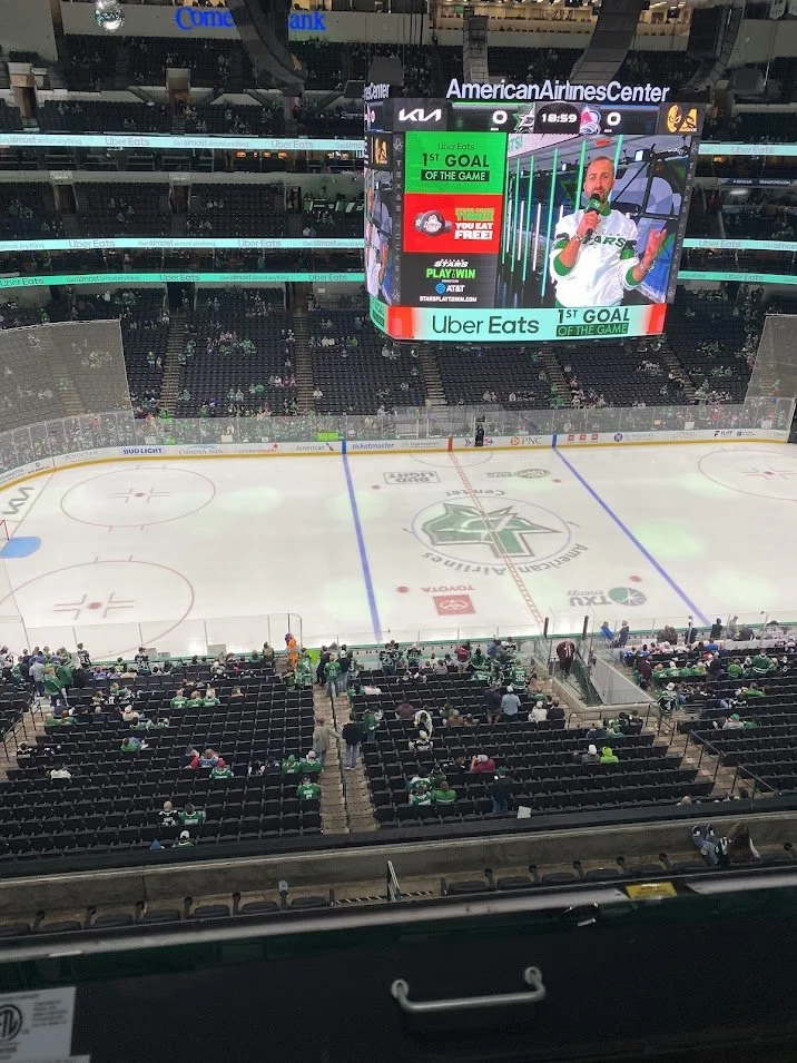 View of an ice hockey rink at American Airlines Center with sparse spectators, some in green jerseys, and a large scoreboard displaying a person speaking during a game.