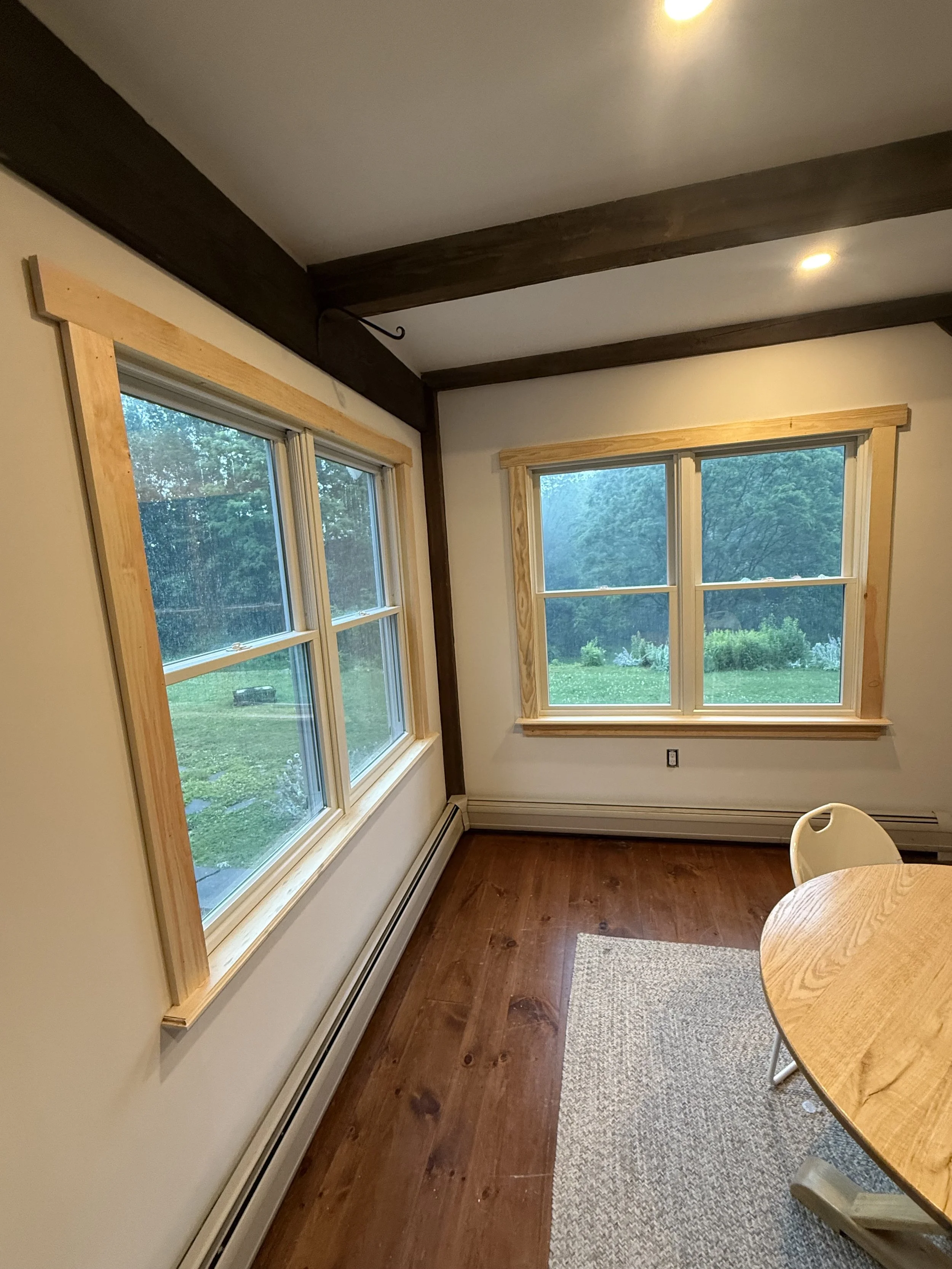 Room with three large windows showing a green outdoor landscape, wooden trim around windows, hardwood floor, a small rug, and part of a round wooden table with a white chair.