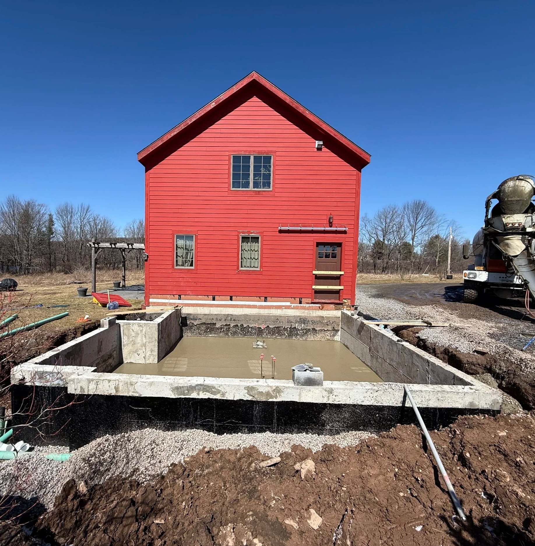 Construction site with a poured concrete foundation for a building, with a red house in the background and construction materials and tools around.
