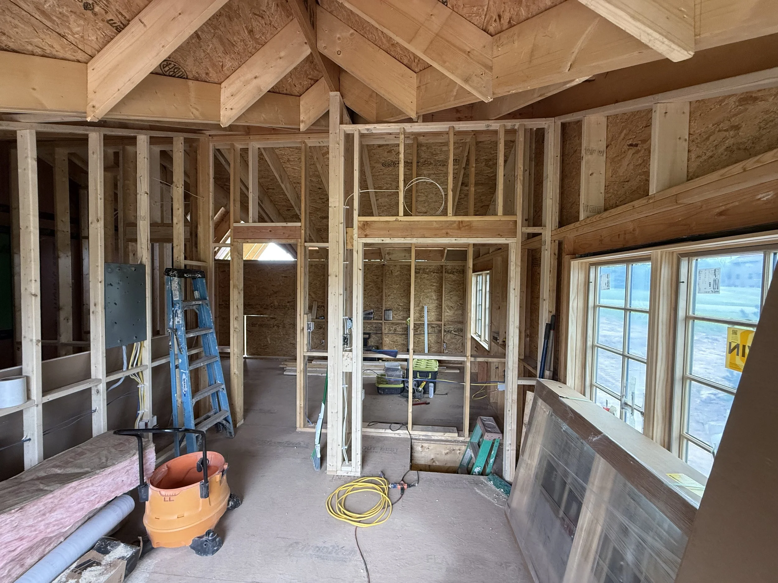 Interior view of a house under construction with exposed wooden framing, windows, construction tools such as a ladder, air compressor, and extension cords seen on a concrete floor.