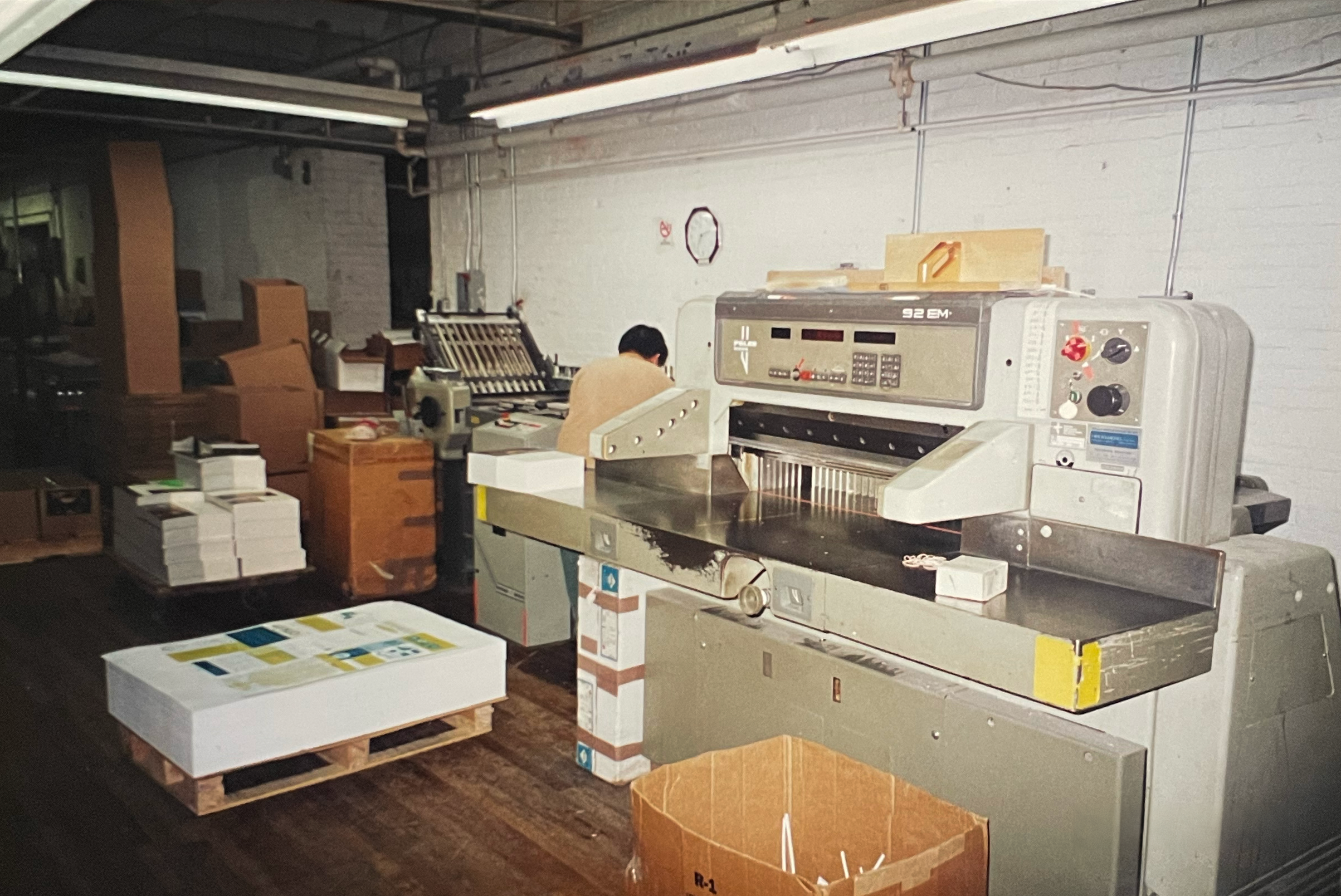 A printing or packaging room with a large industrial machine on the right and a person working in the background, surrounded by stacked boxes and pallets.