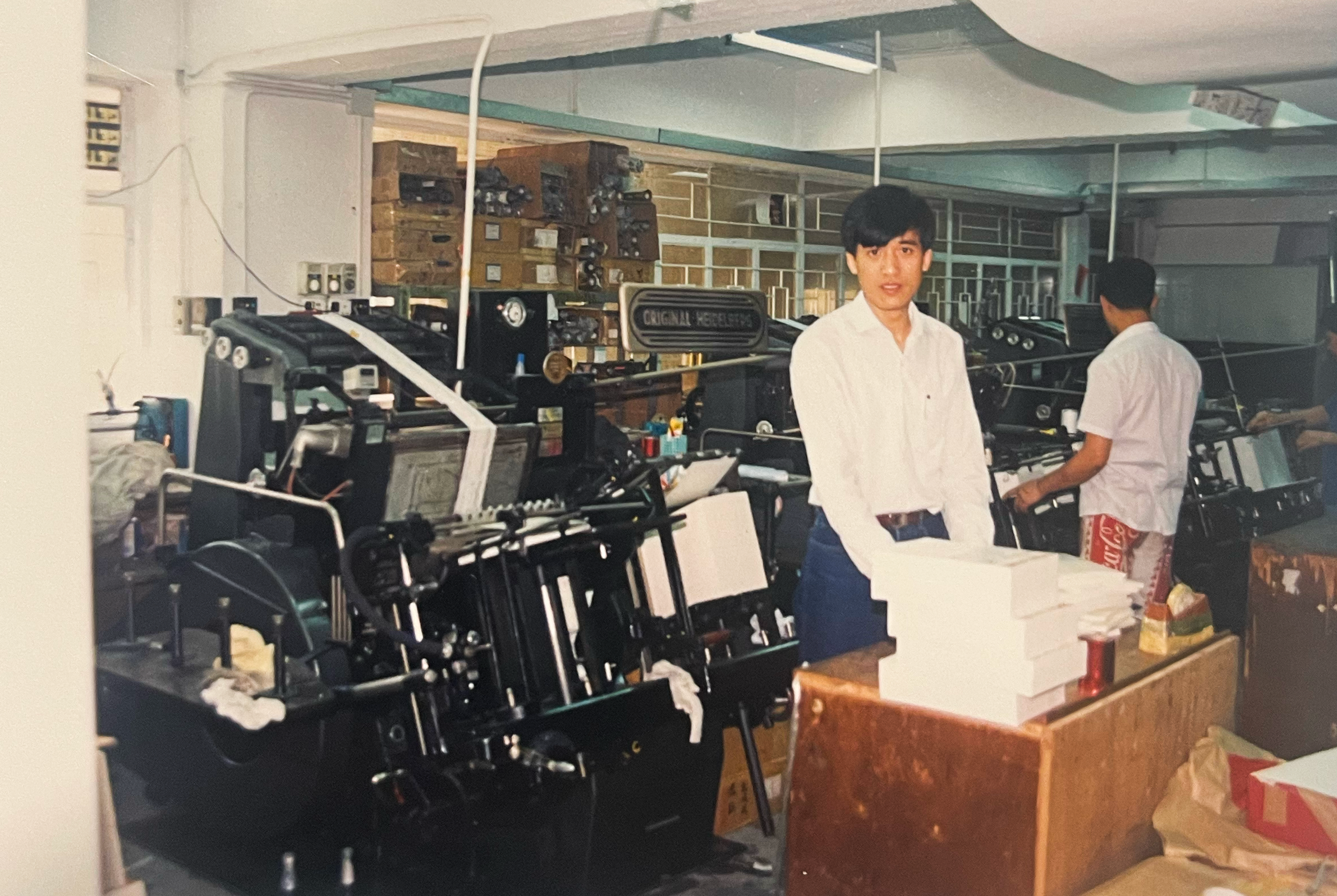 A young man in white shirt and blue jeans stands in a printer workshop next to a large printing machine with stacks of white paper in front of him. Other workers are seen in the background.