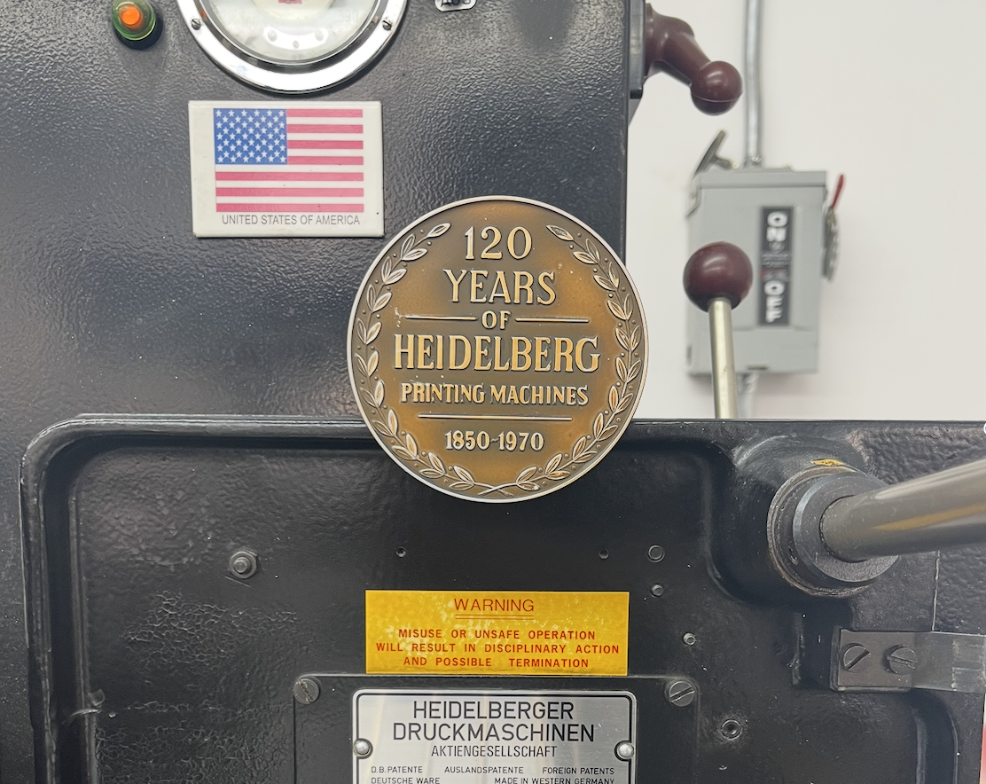 Metal machine with a commemorative plaque celebrating 120 years of Heidelberg printing machines from 1850 to 1970, a warning label, an American flag sticker, and some control levers and dials.