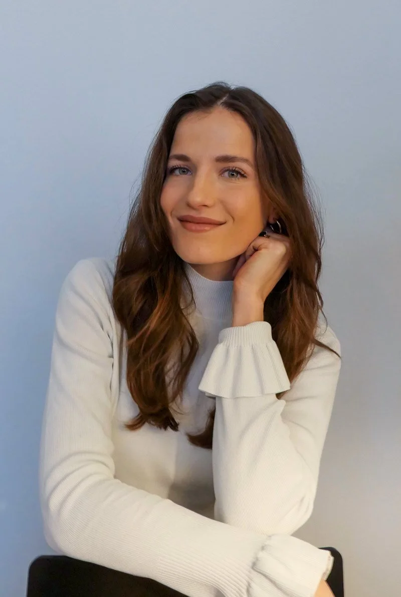 A young woman with long brown hair, wearing a white long-sleeve top with ruffled cuffs, posing with her hand resting on her cheek, smiling softly against a plain light blue background.