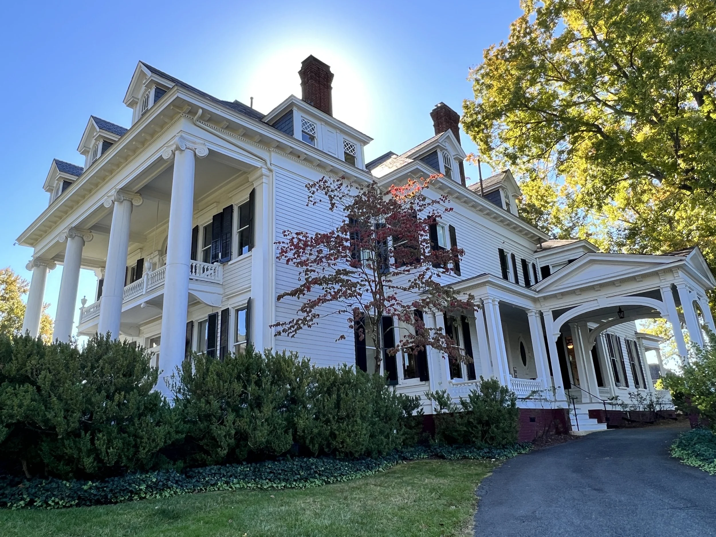 Large white Victorian-style house with tall columns, black shutters, and detailed trim, surrounded by green trees and bushes on a sunny day.