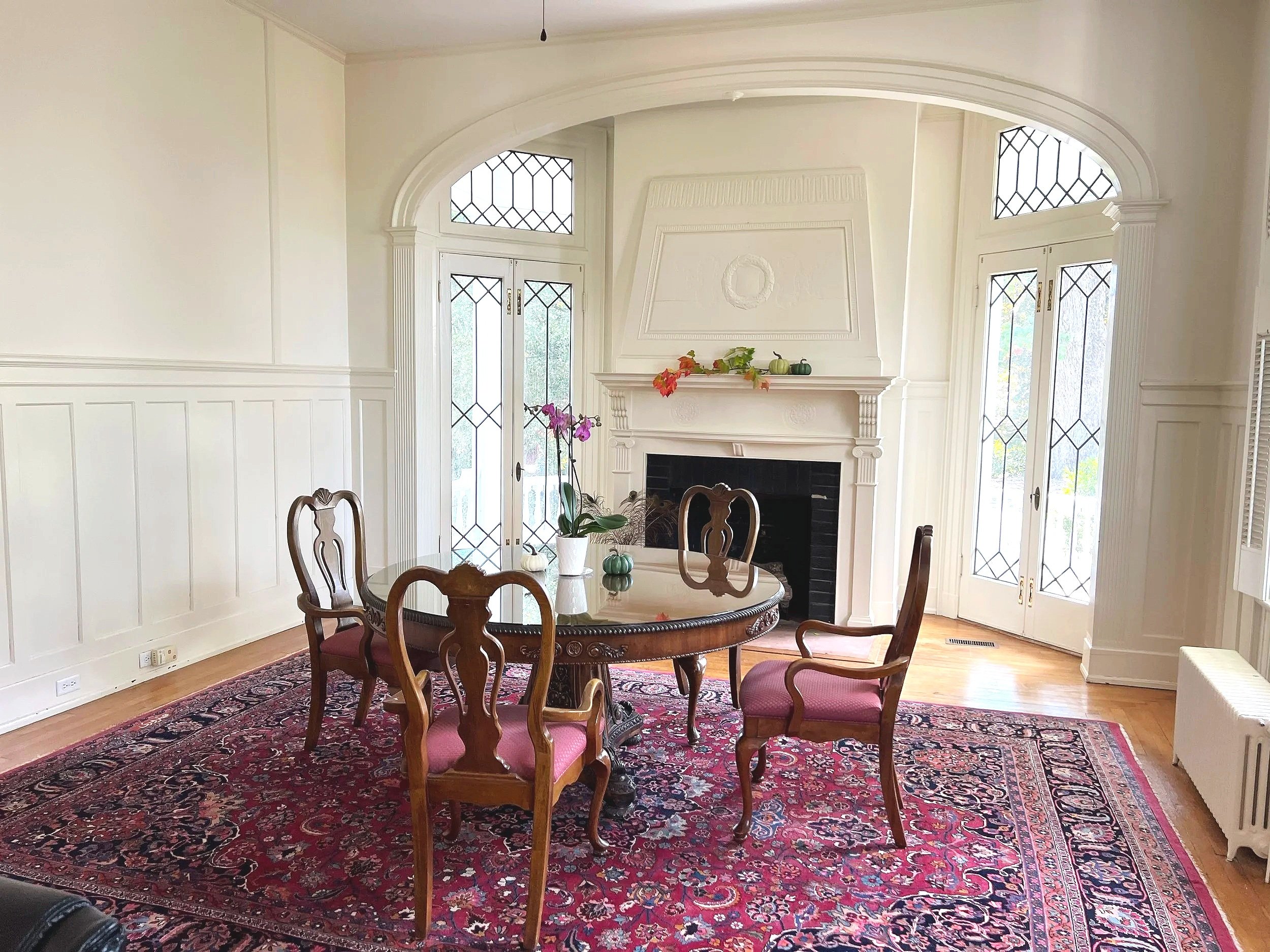 A dining room with a round wooden table and four chairs, a red patterned rug, a white fireplace with decorative mantel and framed wall relief, large windows with diamond pattern glass, and potted plants on the mantel.