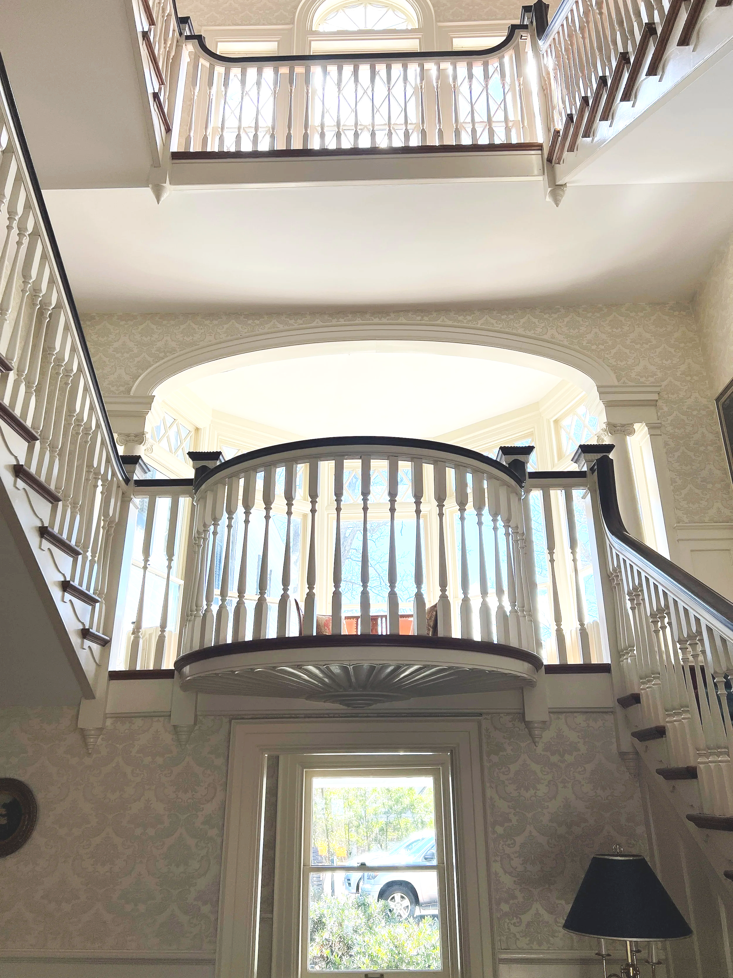 Interior view of a multi-story house with a staircase, featuring a balcony with white balusters and dark wooden handrails, large windows allowing natural light, decorative wallpaper, and a lamp in the bottom right corner.