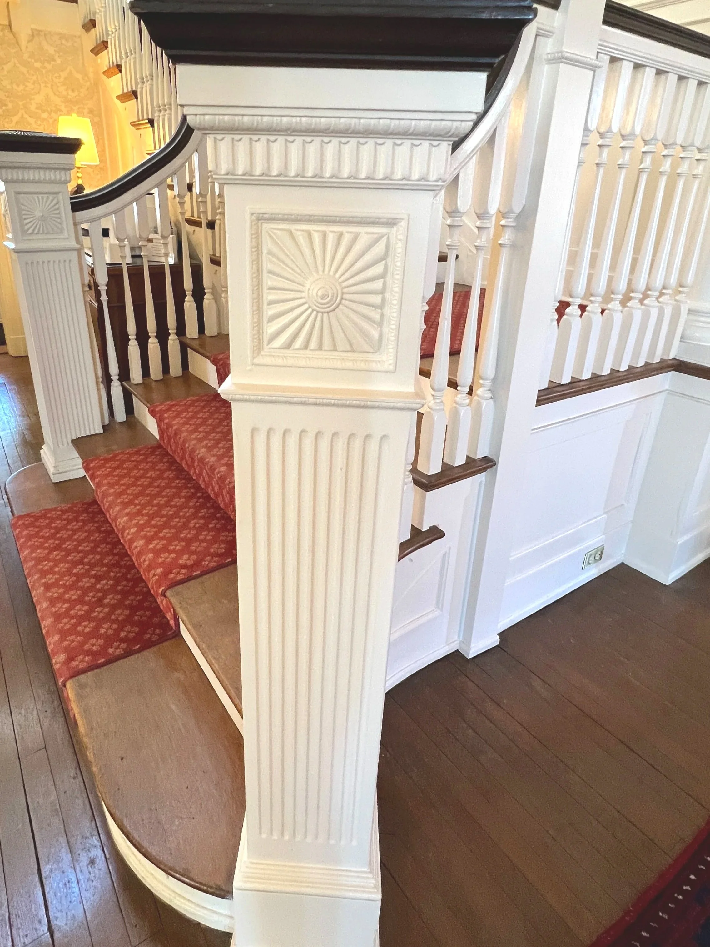 Close-up of a staircase with white wooden balustrade, red patterned stairs, and a dark wooden handrail, inside a house with ornate wallpaper and warm lighting.