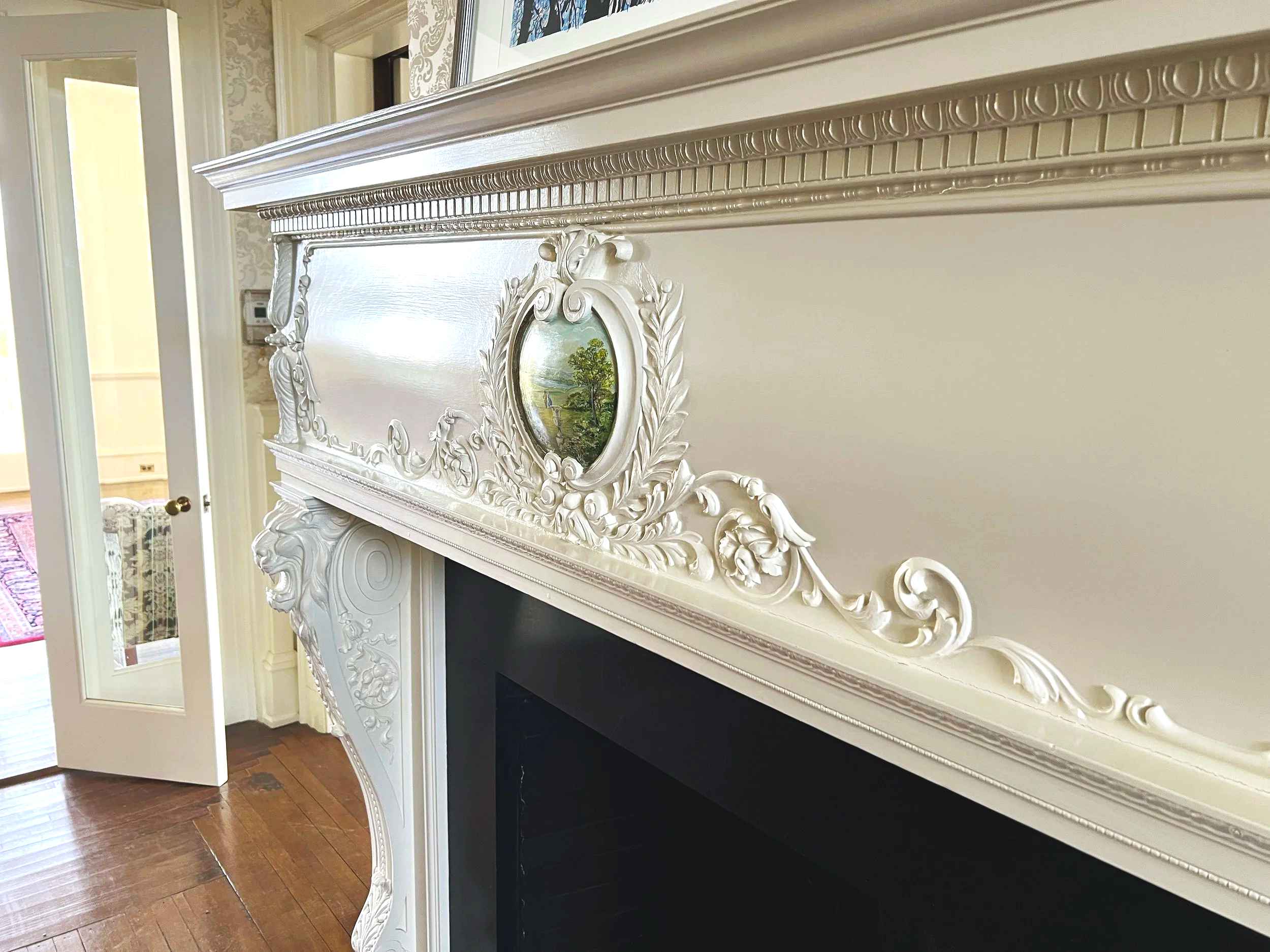 Close-up of an ornate white fireplace mantel with detailed carvings and a small oval landscape painting in the center.