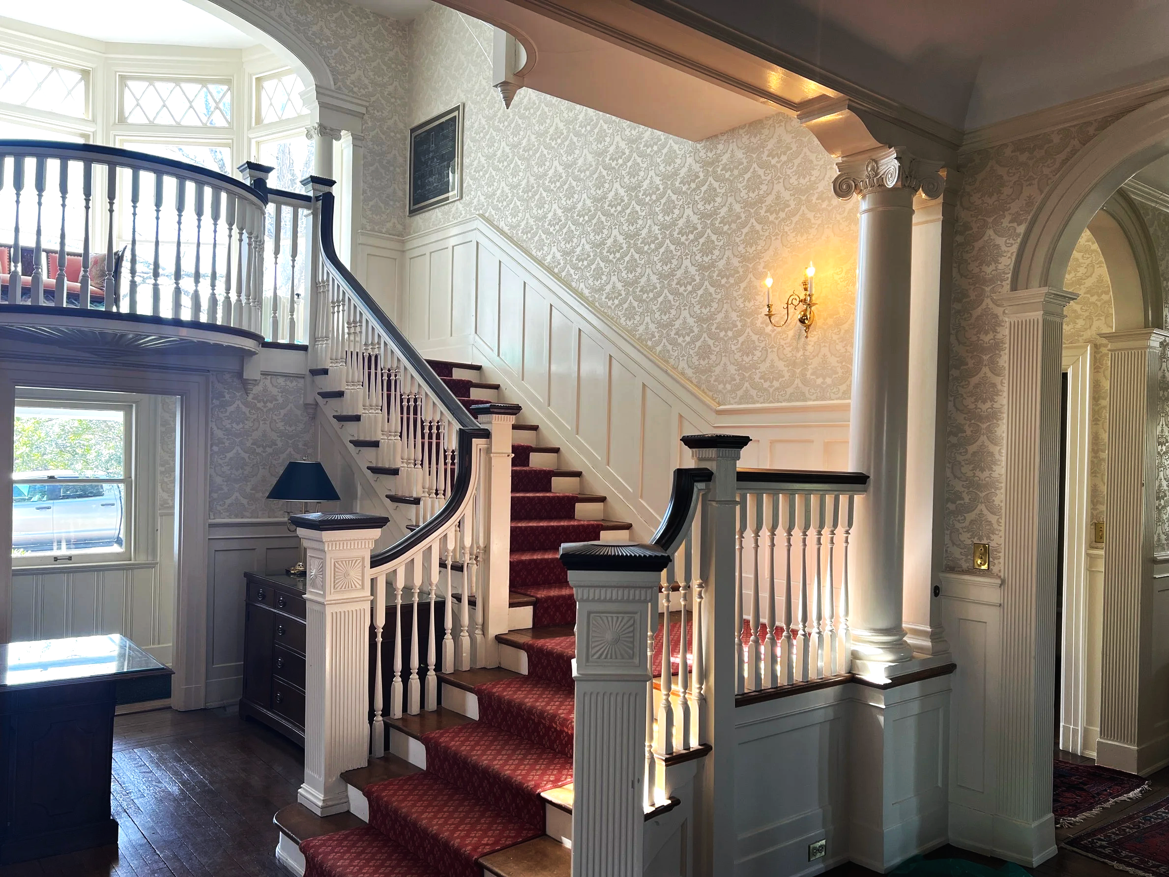 Elegant staircase with red carpet and white banisters leading to an upper floor in a Victorian-style home with patterned wallpaper, wall sconces, and wooden flooring.