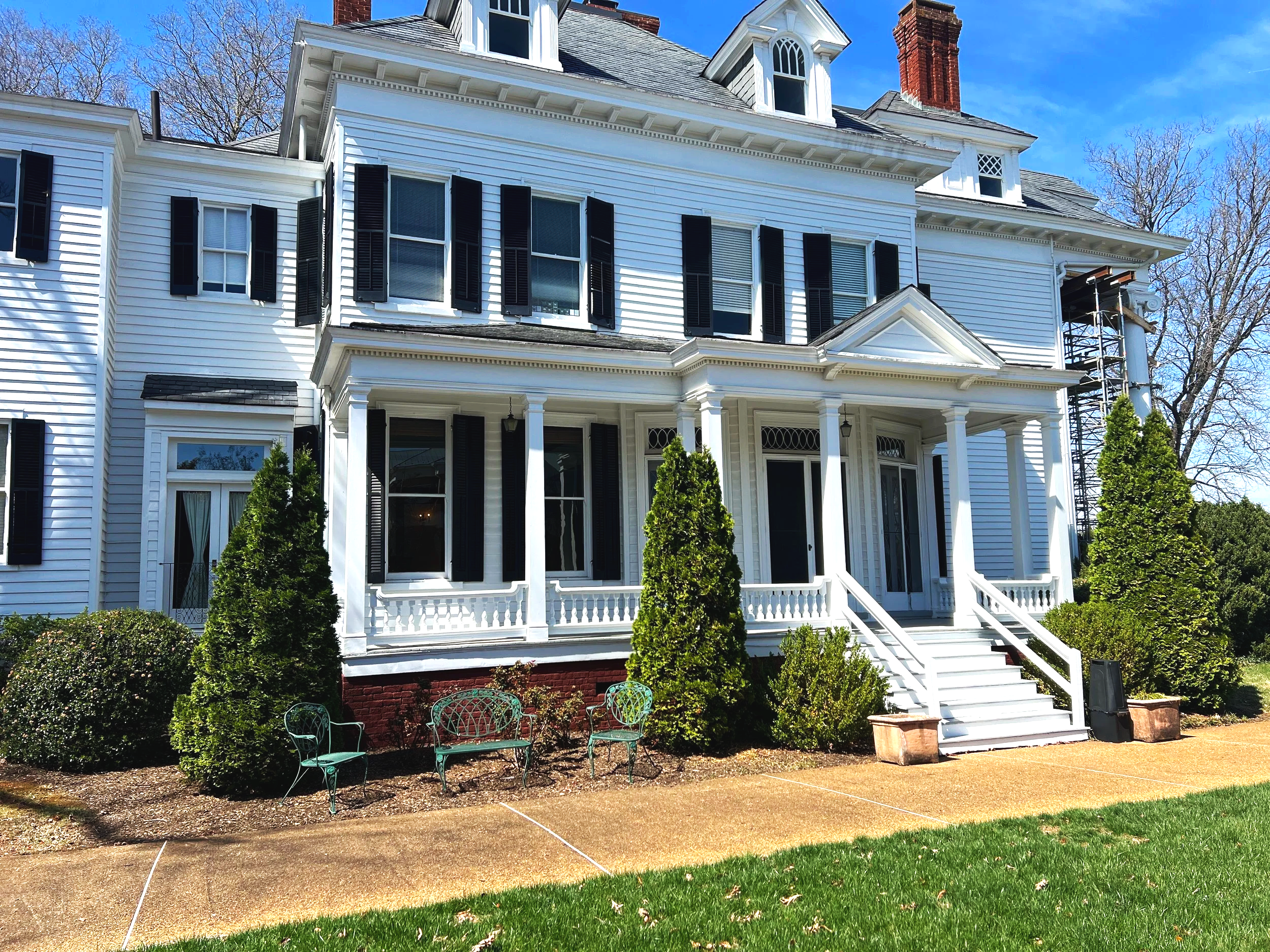 A large white Victorian-style house with black shutters, a front porch with columns, and a staircase leading to the front door. There are green bushes and trees in front, with a concrete walkway and a grassy lawn.