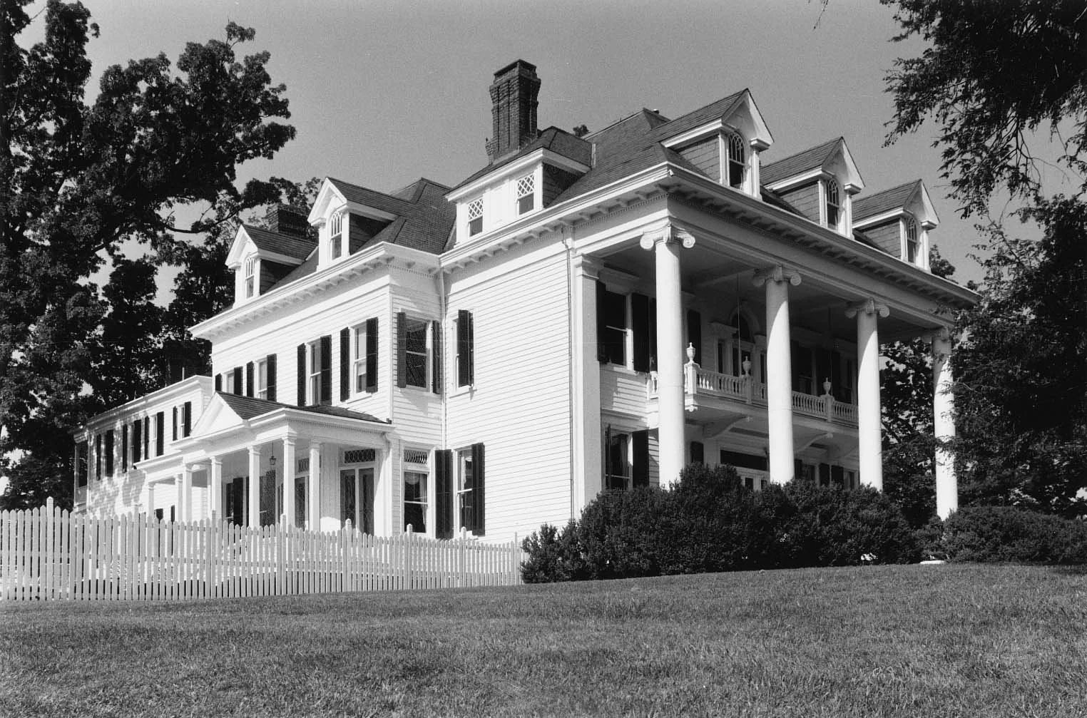 Black and white photo of a large, elegant, Victorian-style house with multiple stories, a covered porch supported by white columns, and dormer windows on the roof.
