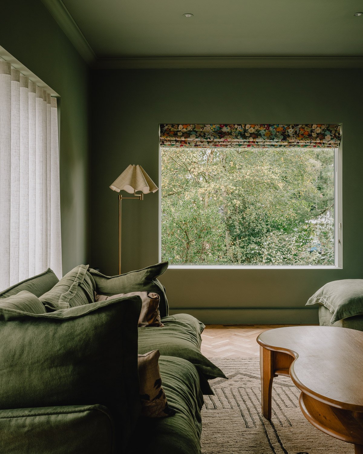 Living room with green walls, a large window overlooking trees, a green sofa with cushions, a standing lamp, and a wooden coffee table.