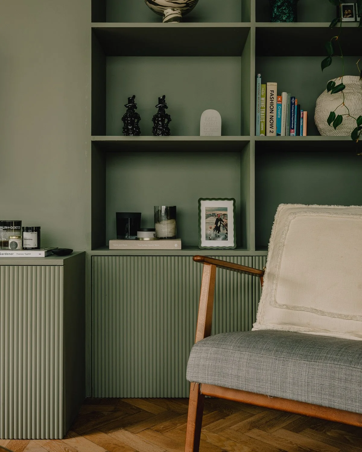Living room with green shelving unit, decorative items, books, a framed photo, and a beige armchair with wooden armrests.
