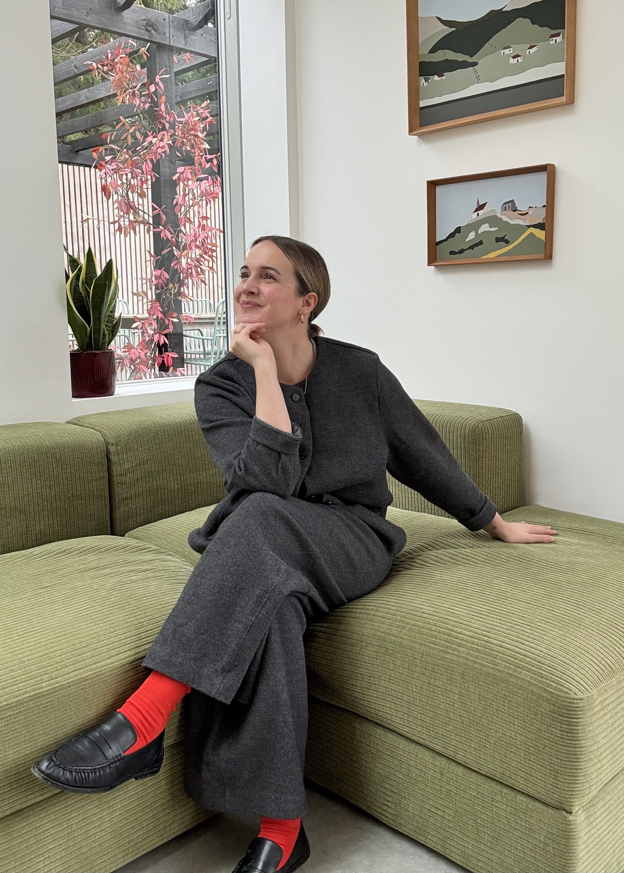 A woman sitting on a green couch in a room with white walls, decorated with two landscape paintings. She is wearing a dark gray outfit with red socks and black loafers, and is looking thoughtfully out the window near a potted plant with pink and green foliage.