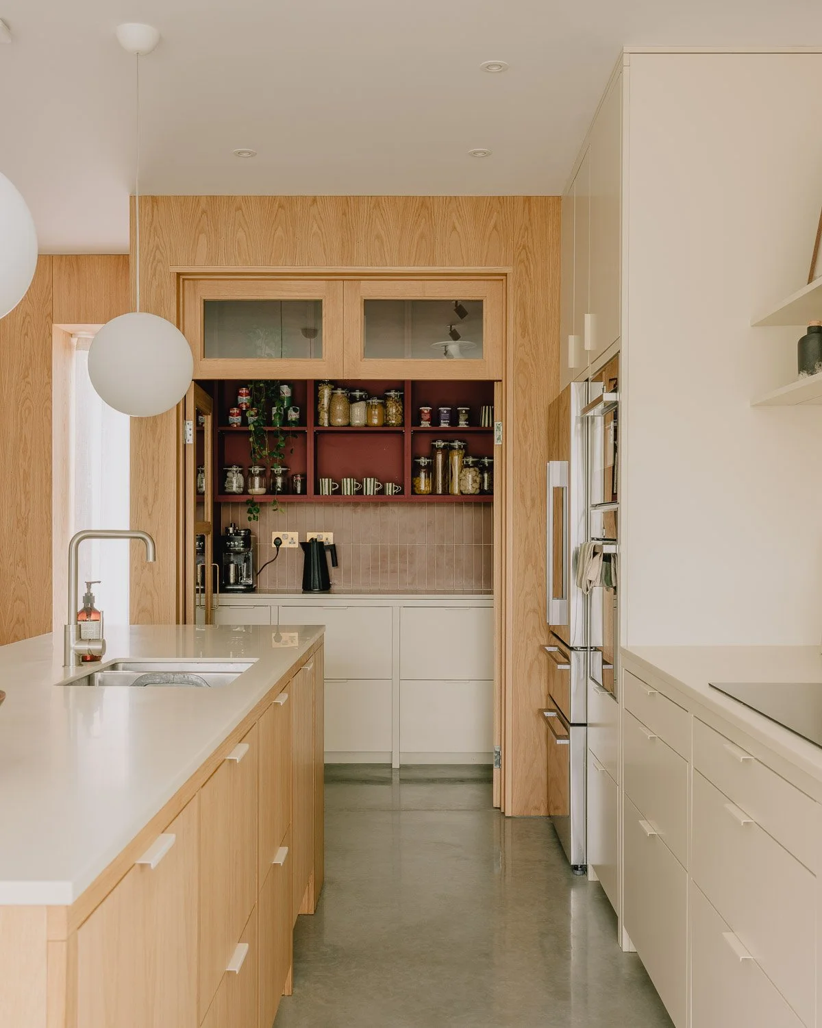 Modern kitchen with white countertops, wooden cabinets, and open shelves filled with jars and dishes, featuring a stainless steel refrigerator and built-in ovens.