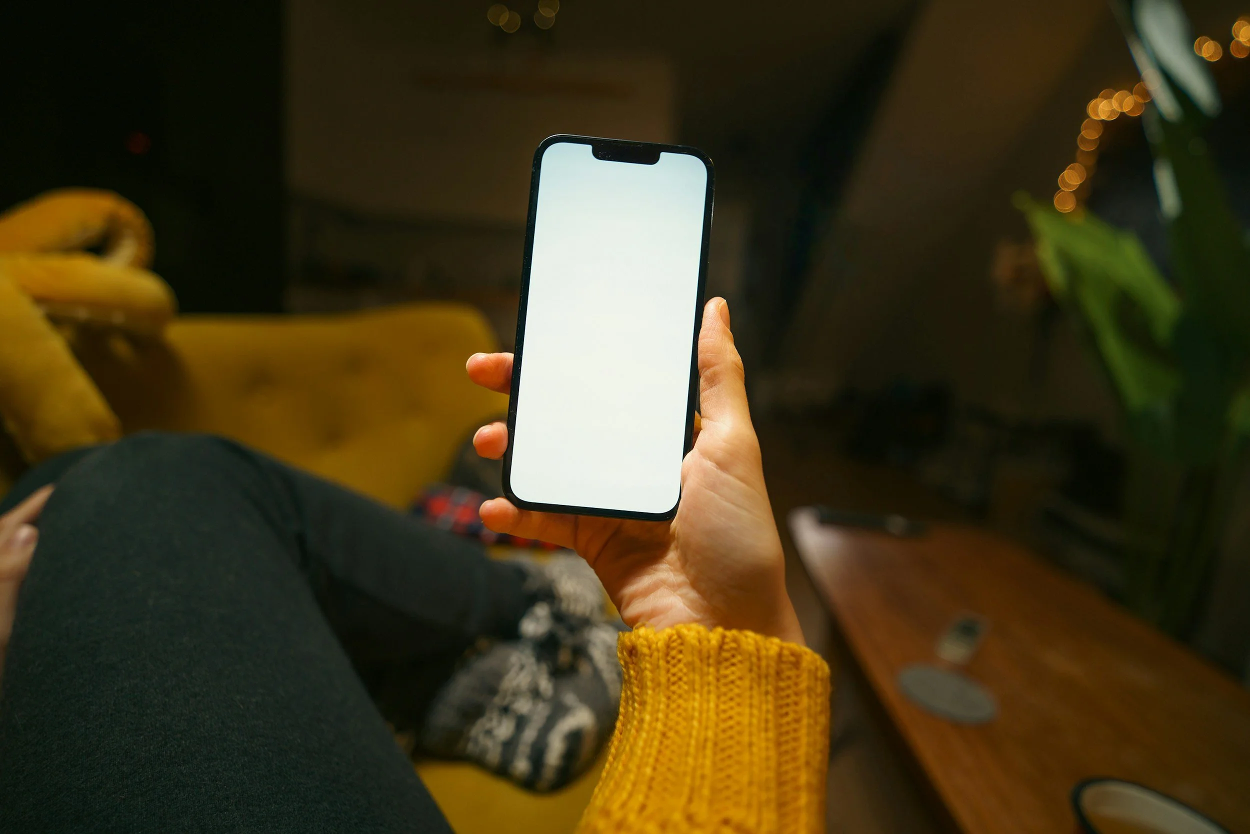 A person holding a smartphone with a blank white screen, sitting on a yellow sofa in a dimly lit room decorated with string lights and green plants.
