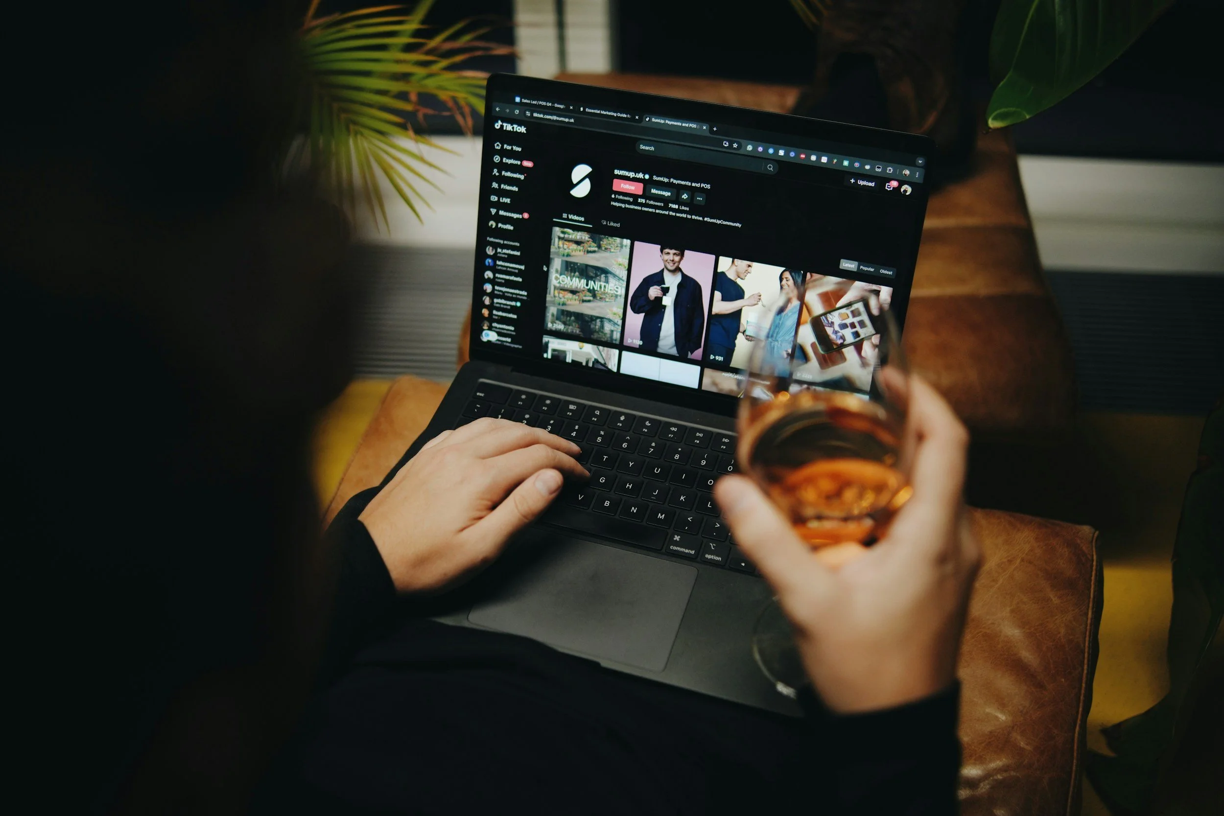 A person using a laptop and holding a glass of wine, browsing TikTok on a dark screen with a background of plants and a leather surface.