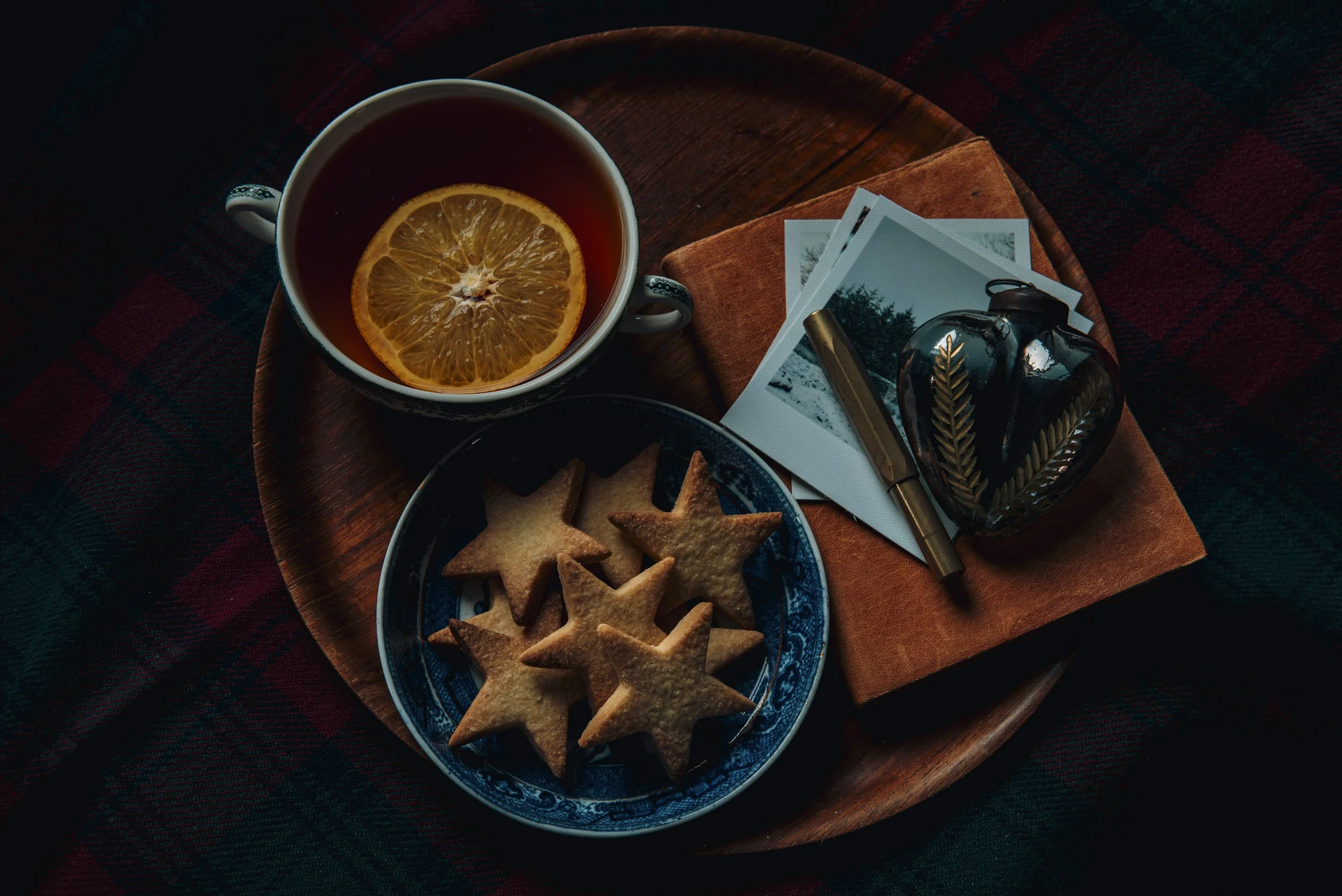 A cup of tea with a lemon slice, a plate of star-shaped cookies, several photographs, a pen, and a decorative glass bottle on a wooden tray.