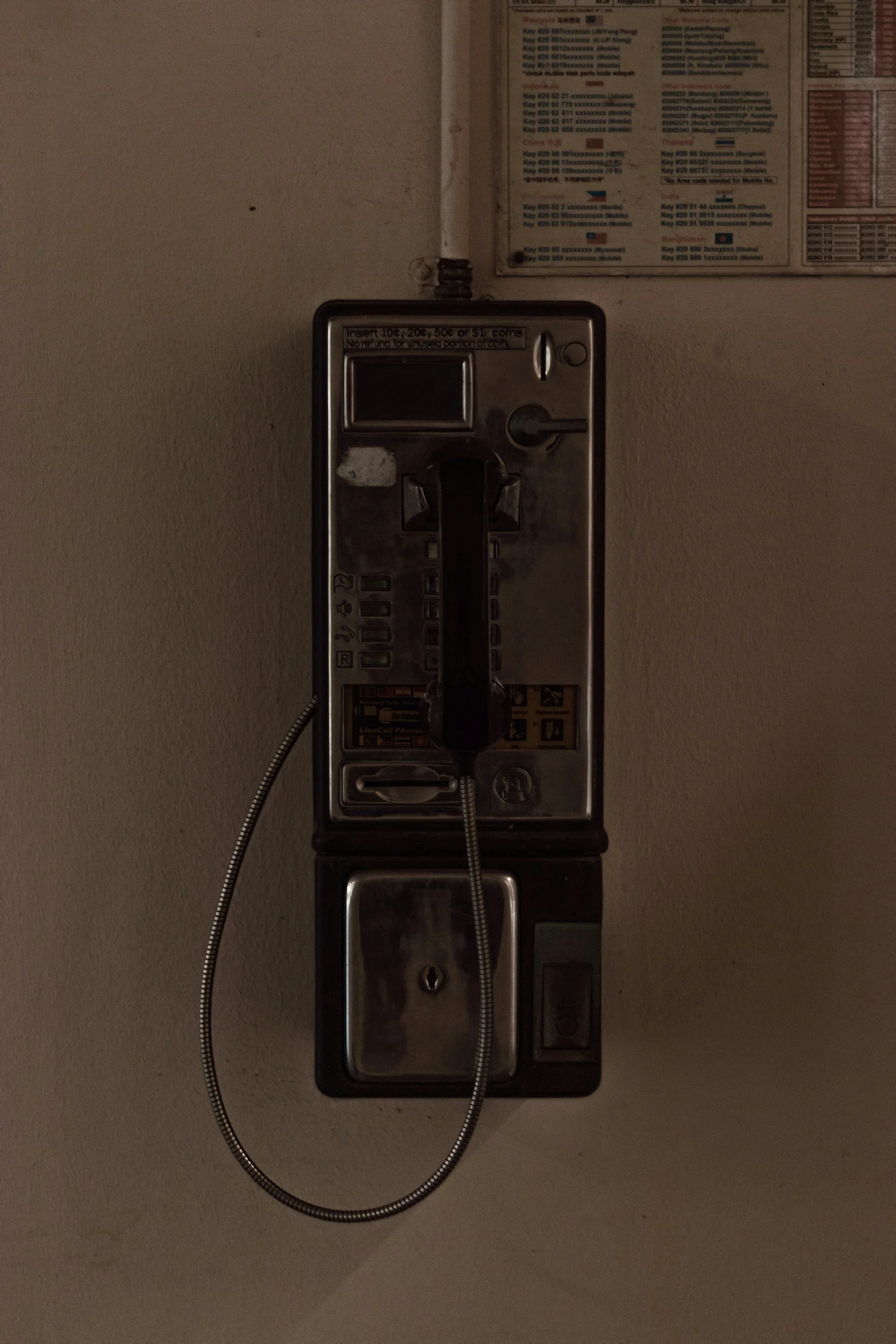 An old payphone mounted on a beige wall, with a payphone card slot, keypad, and coin slot, along with a mounted phone receiver and a keypad below.