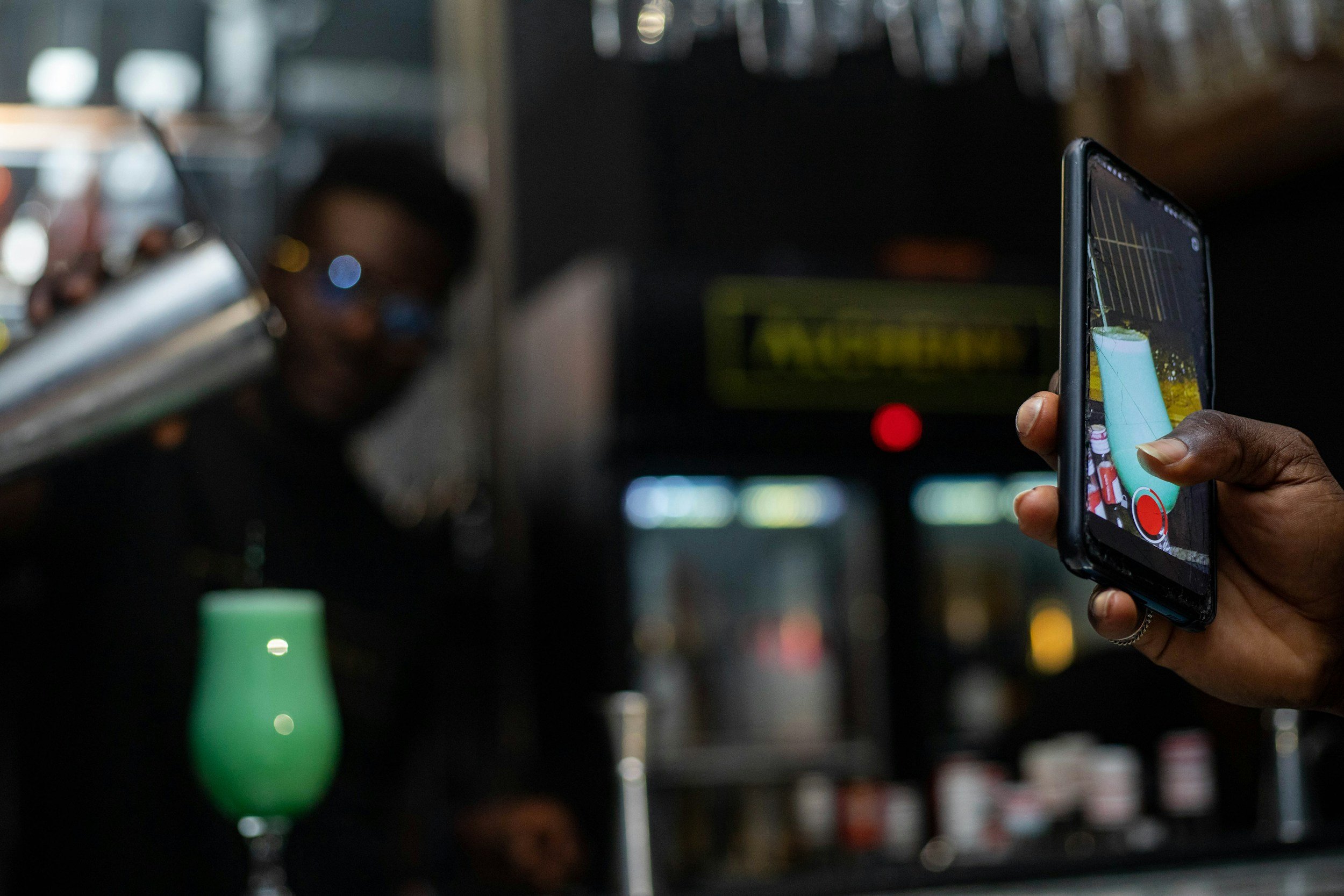 Person recording a bartender pouring a drink with a smartphone in a bar setting.