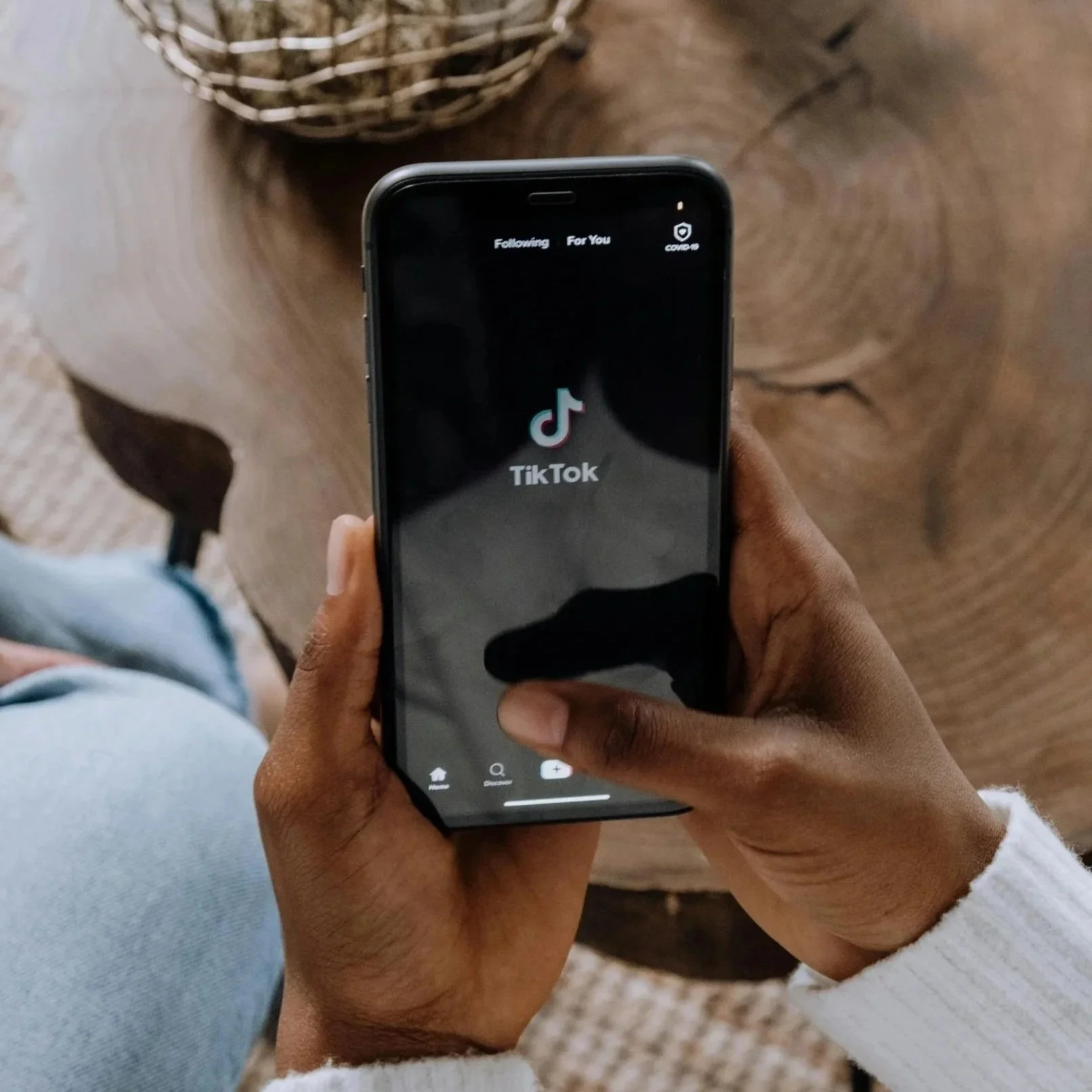 Person holding a smartphone displaying the TikTok app logo on the screen, sitting at a wooden table.