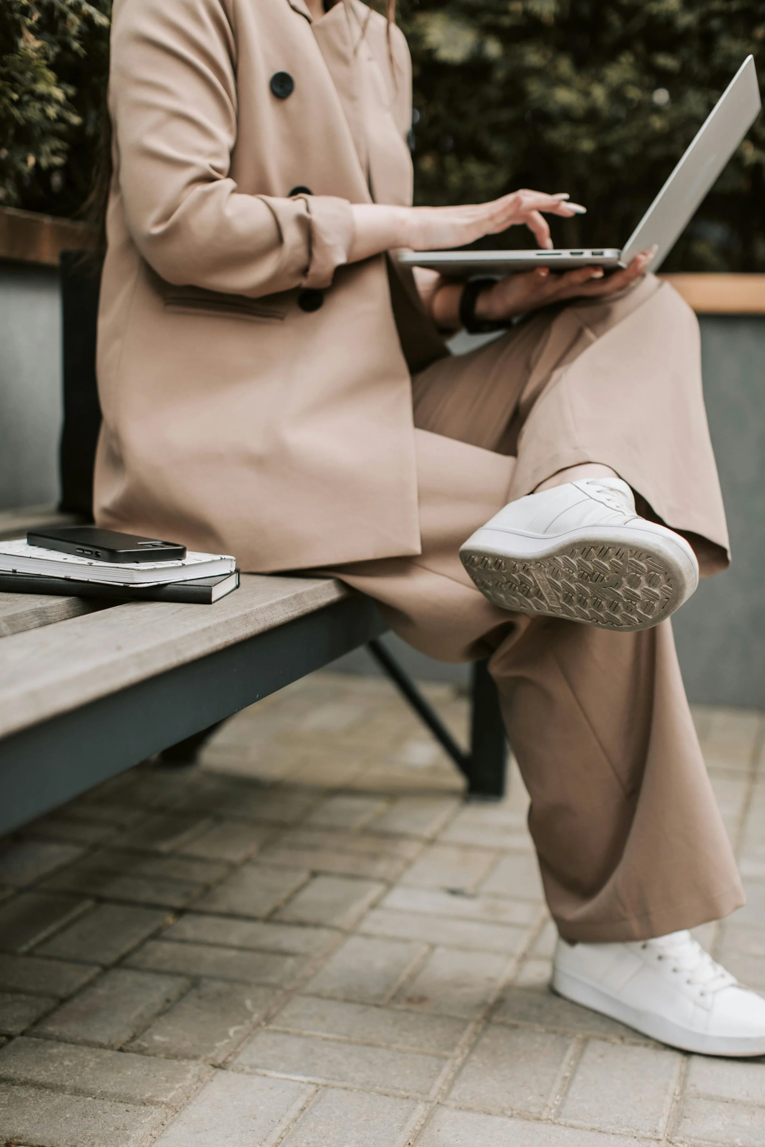 Person sitting on a bench using a laptop outdoors with notebooks and a phone nearby.