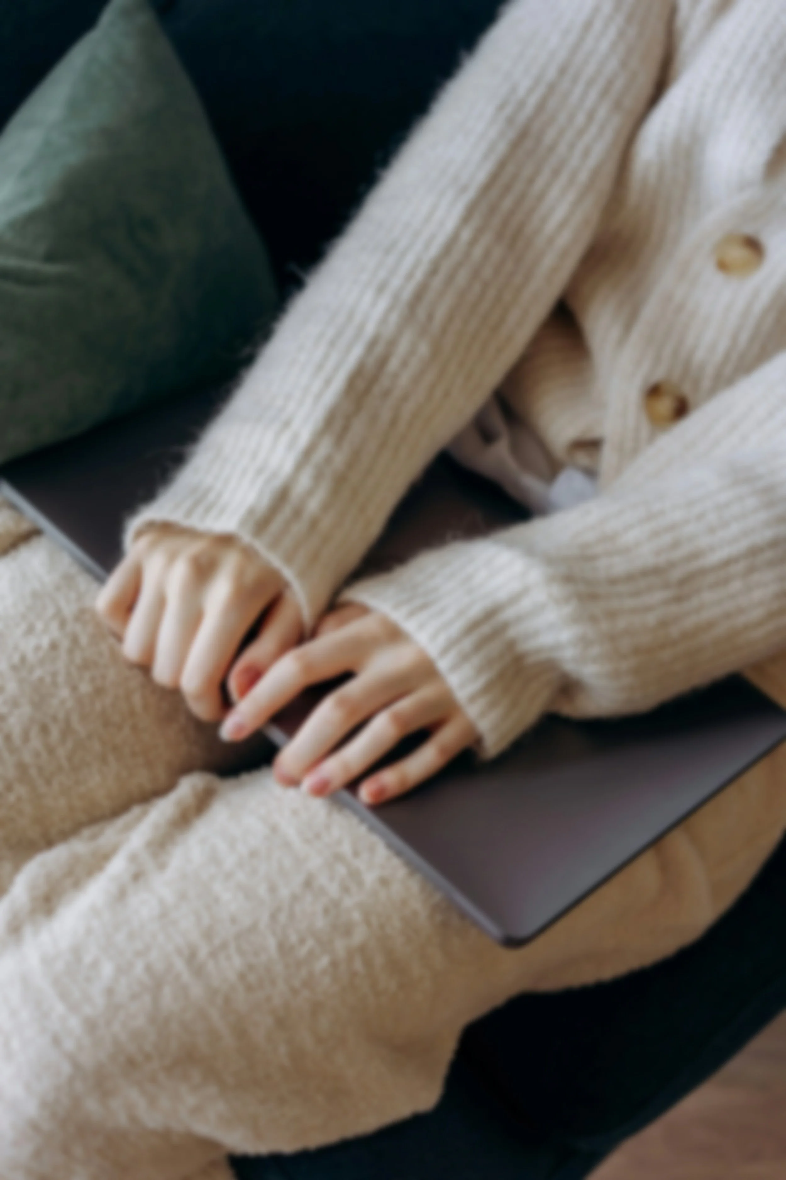 Person wearing a cream-colored knit sweater and beige pants working on a laptop on a beige couch.