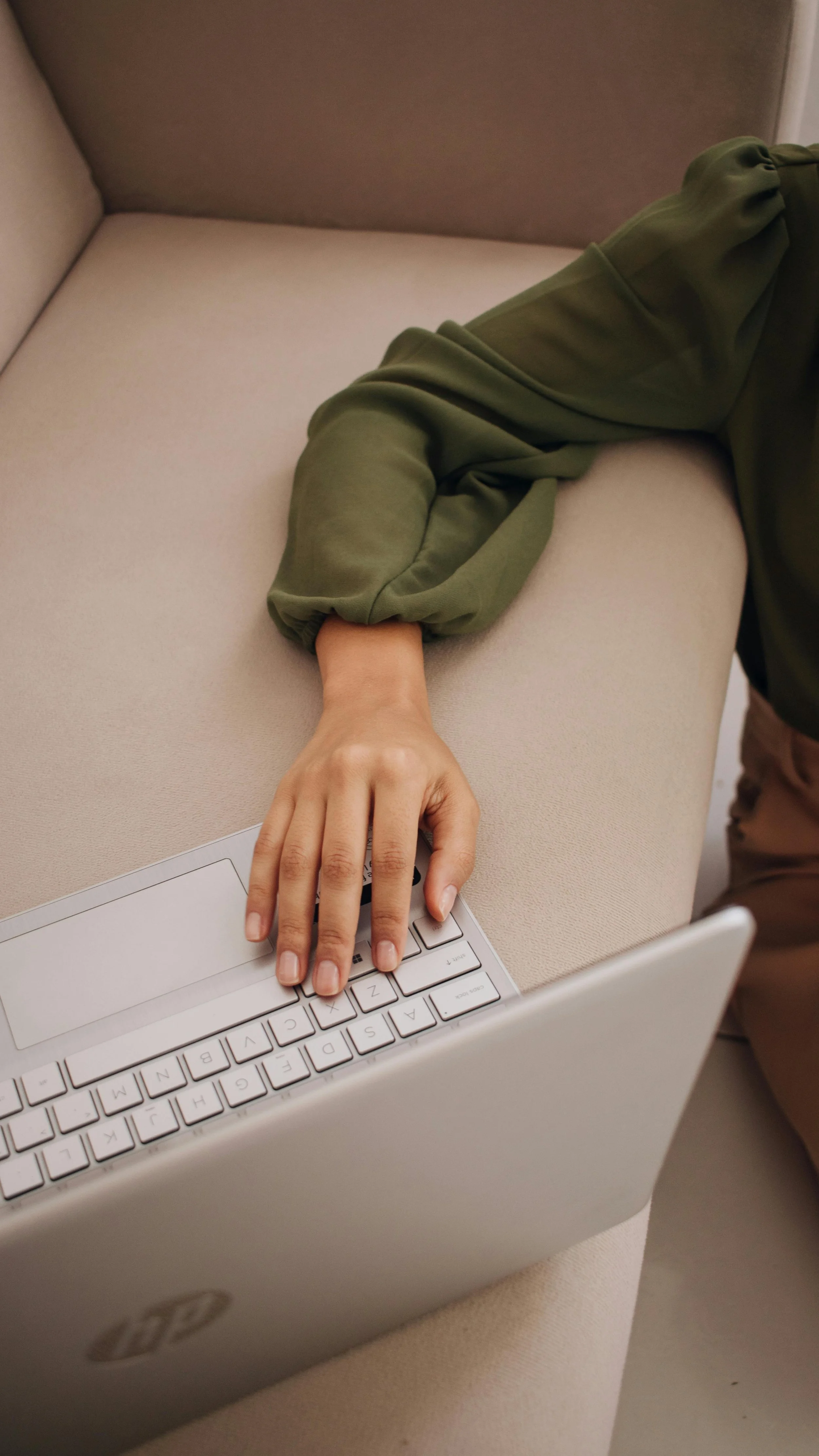 Person sitting on a beige couch using a silver laptop, with their hand on the keyboard and wearing an olive green sweatshirt.
