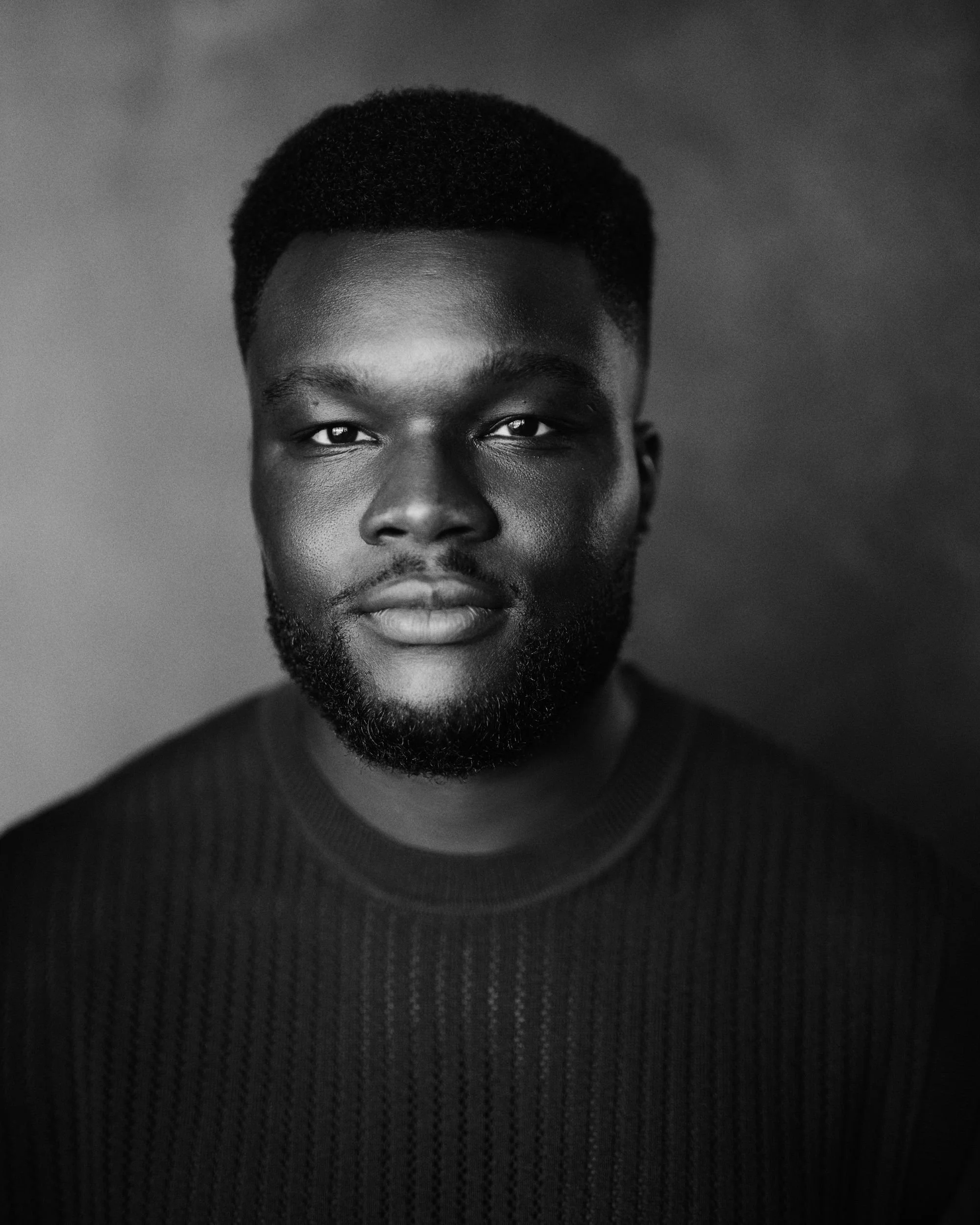 Black and white close-up portrait of a young man with short curly hair and beard, wearing a ribbed dark sweater, facing directly at the camera.