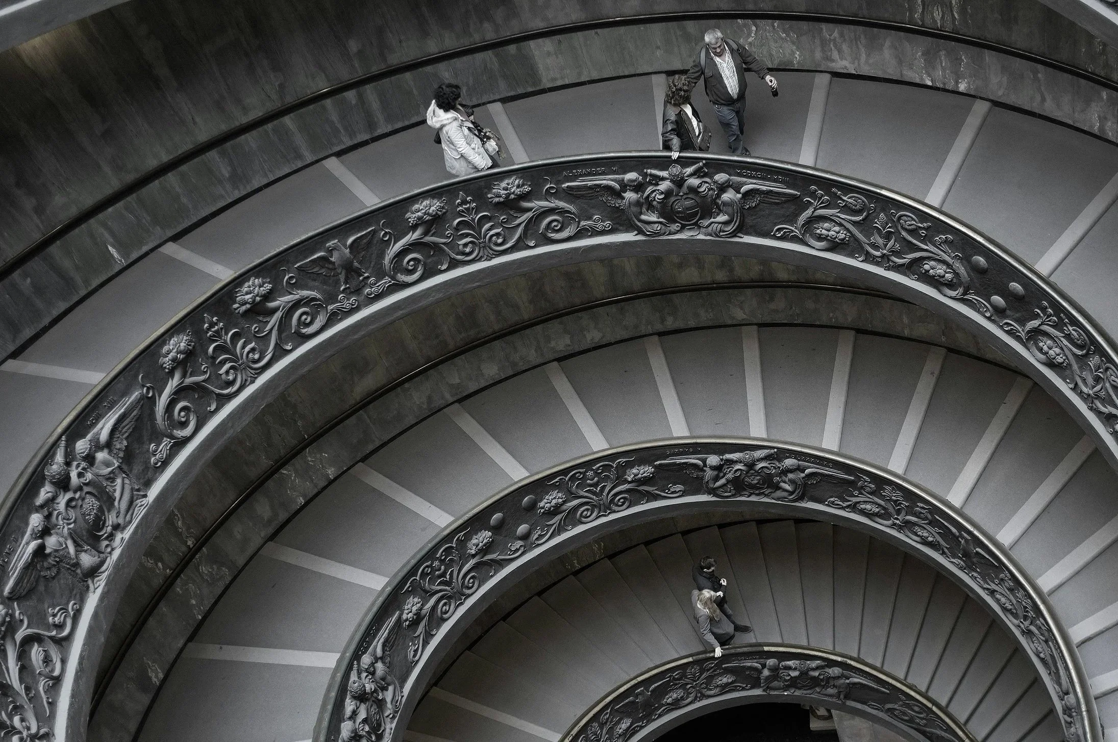 A spiral staircase viewed from above, with intricate decorative railings and three people walking down the stairs.