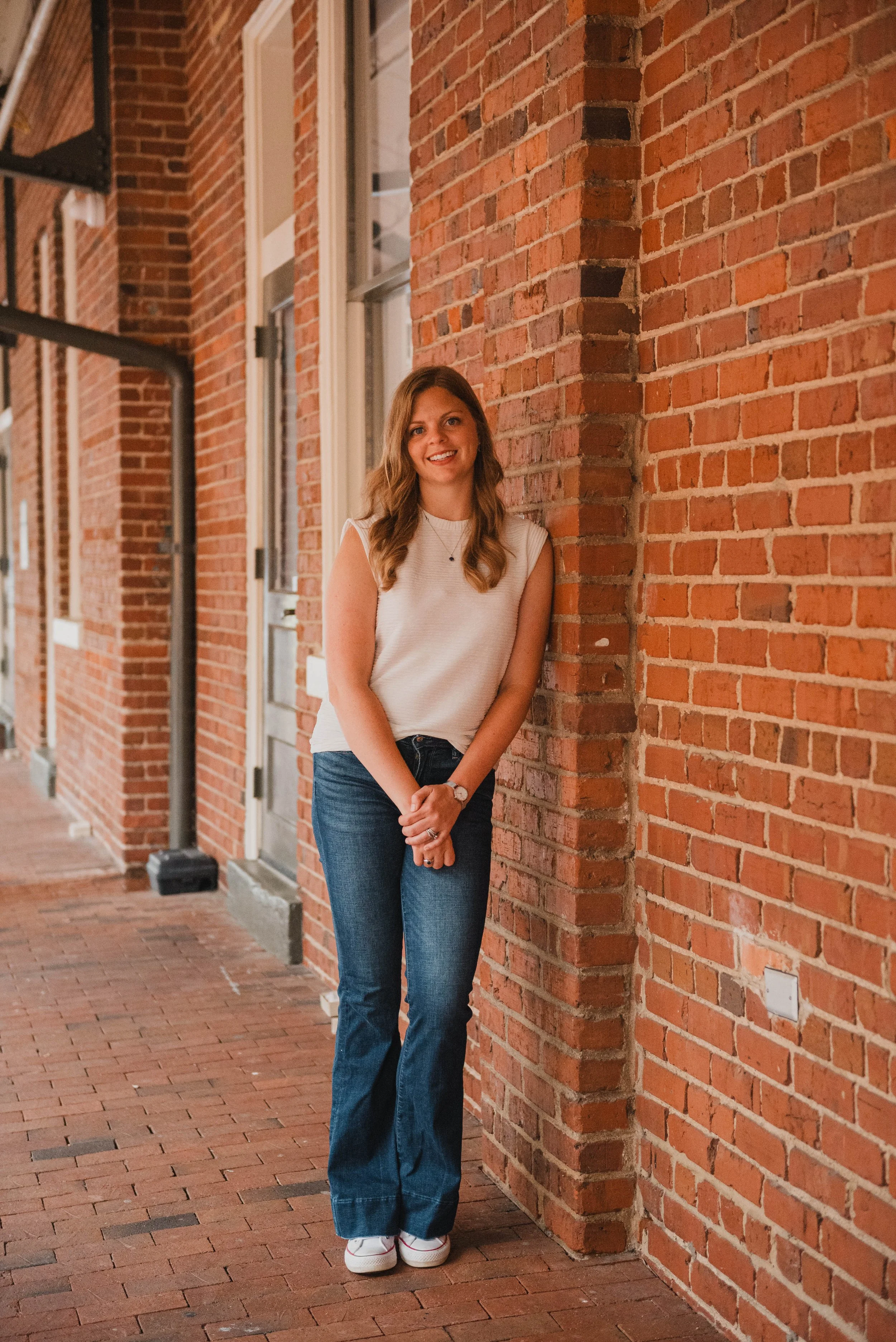 A smiling young woman with wavy brown hair standing against a brick wall on a sidewalk, wearing a sleeveless white top, blue jeans, and white sneakers.