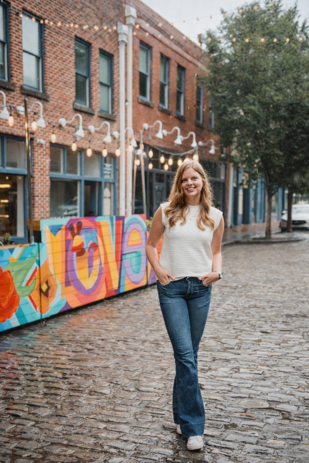 A young woman with long wavy hair smiling and walking on a cobblestone street in an urban area. She is wearing a sleeveless white top, blue jeans, and white shoes. There is a colorful mural with the word 'LOVE' on a barrier to her left, with string lights hanging above and trees lining the street.