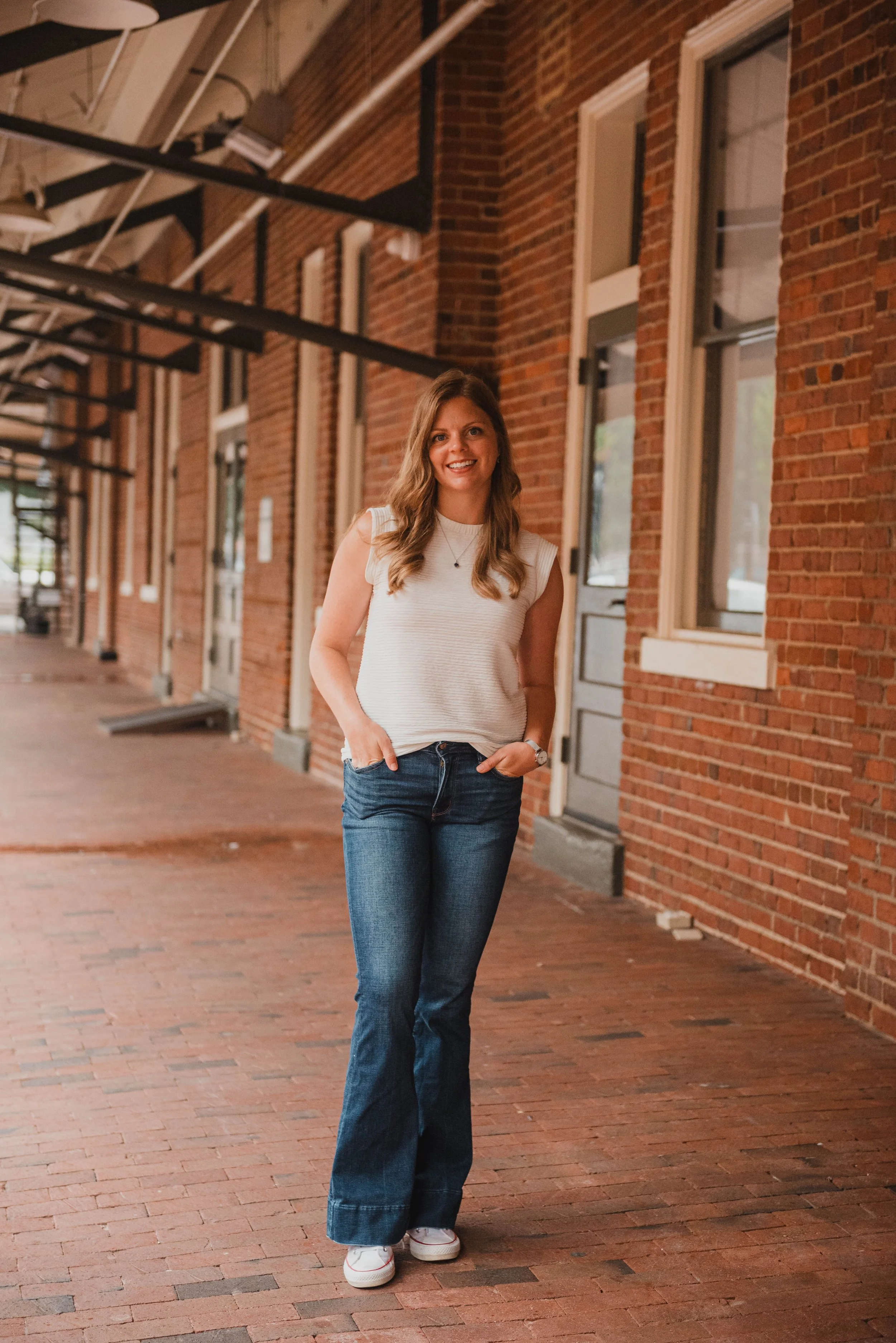 A woman standing on a brick sidewalk next to a brick building with multiple windows, smiling at the camera in a casual, sleeveless white top and jeans.