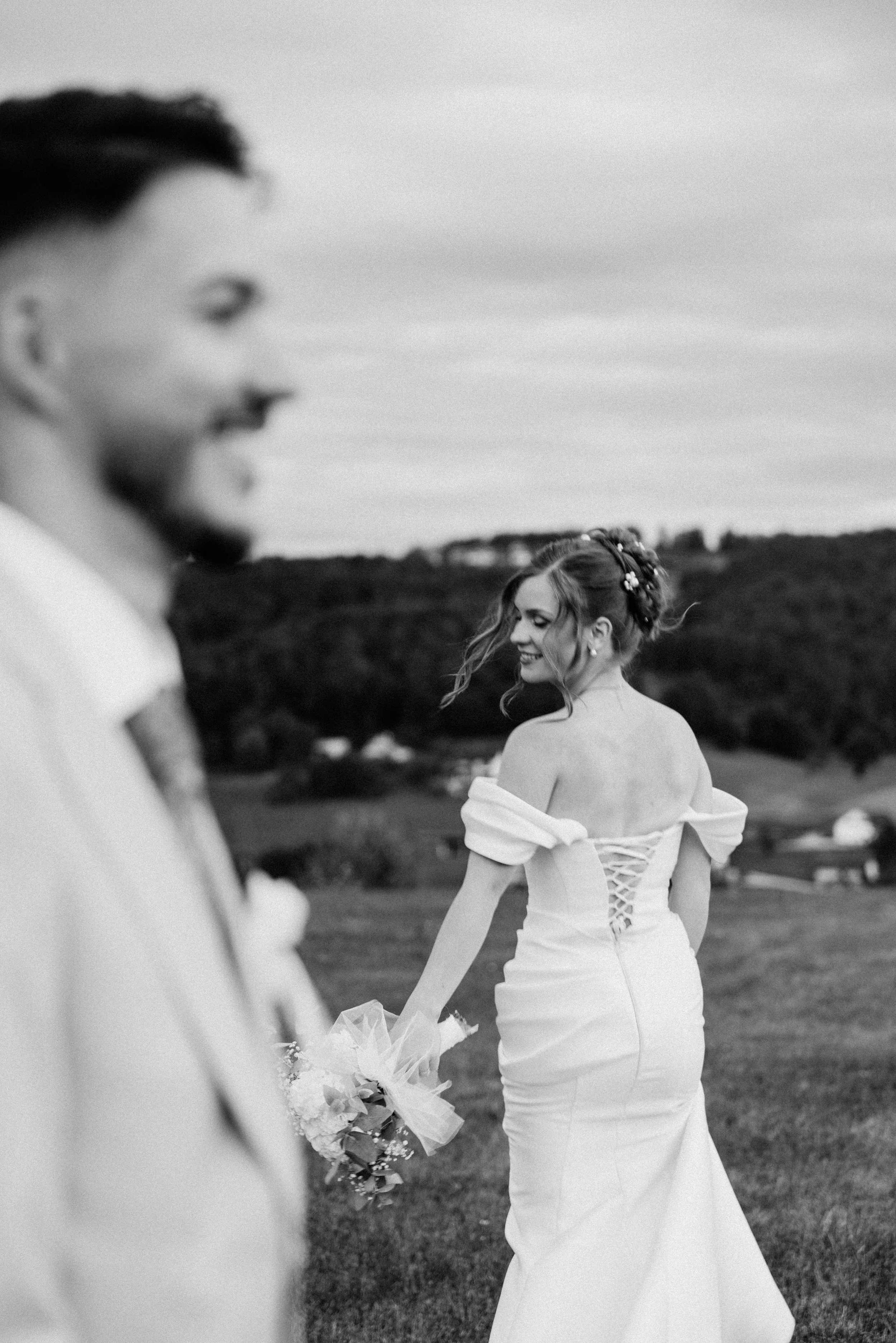 Črno-bela fotografija neveste, ki se ozira proti ženinu s poročnim šopkom, z ženinom v ospredju.