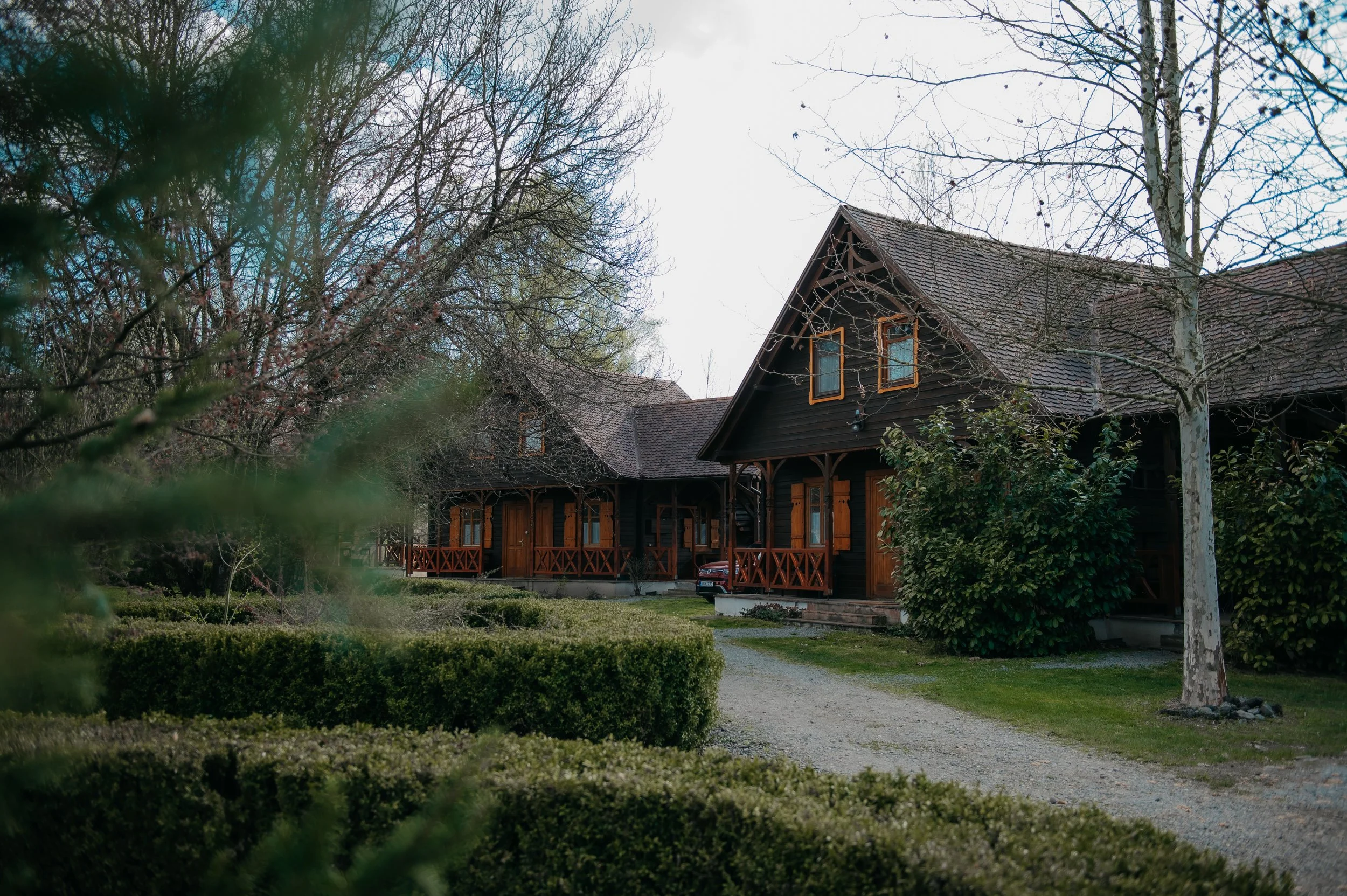 A brown wooden house with a steep roof, surrounded by trees and greenery, under a cloudy sky.