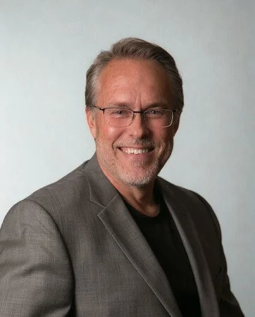 Portrait of a middle-aged man with gray hair and glasses, smiling, dressed in a gray blazer and black shirt, against a light background.