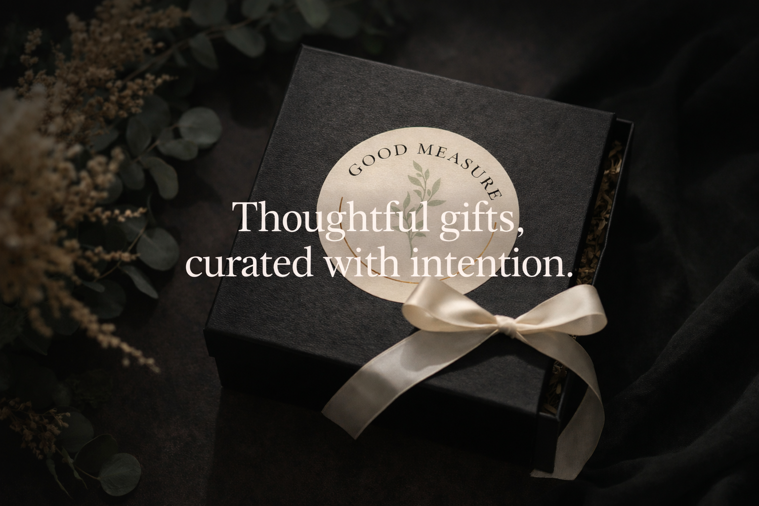 A black gift box with a beige ribbon and a circular label reading "Good Measure" with a plant illustration, surrounded by greenery and dried flowers, with overlaid text saying "Thoughtful gifts, curated with intention."