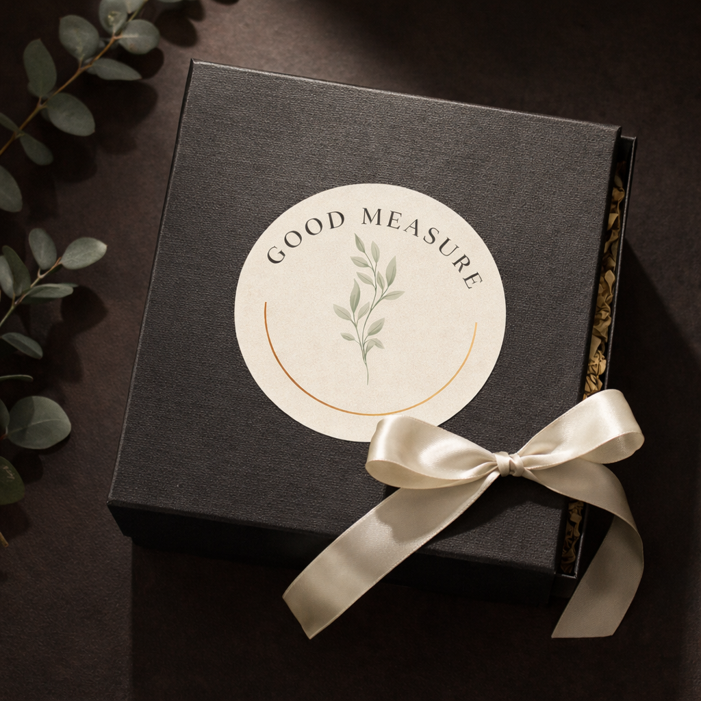 Black gift box with a round label reading 'Good Measure' and an illustration of green leafy plant, tied with a cream satin ribbon, on a dark surface with eucalyptus leaves.