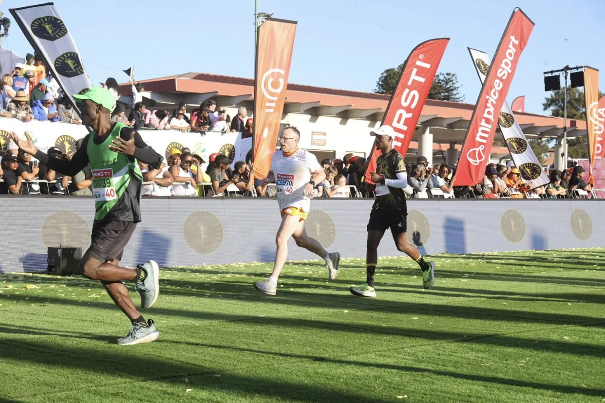 Comrades Marathon runners competing in a race on a grassy field with spectators and flags in the background.