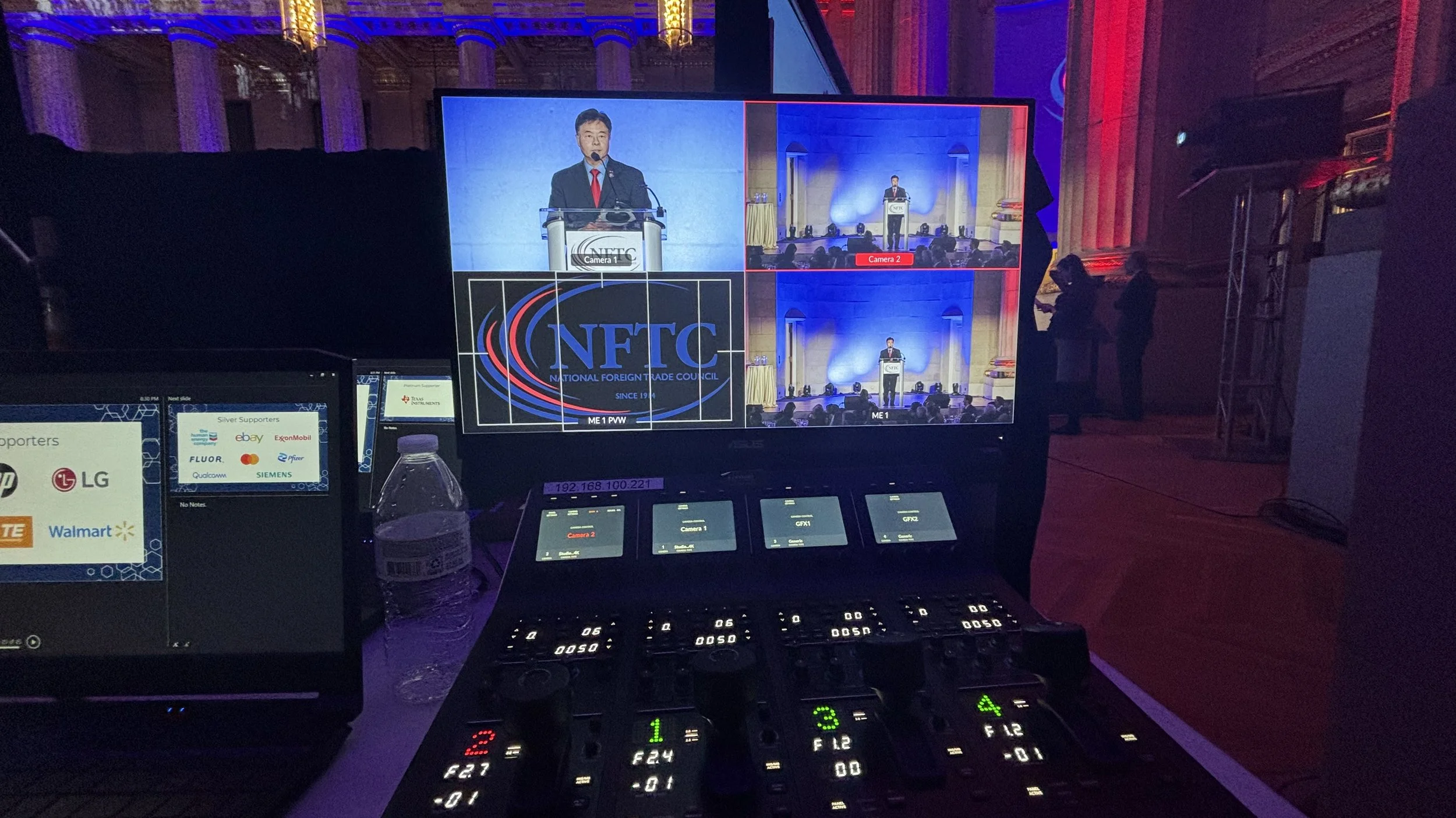 A photo of a conference stage with a man speaking at a podium, projected on multiple screens. The setting includes professional equipment and a control panel in the foreground.