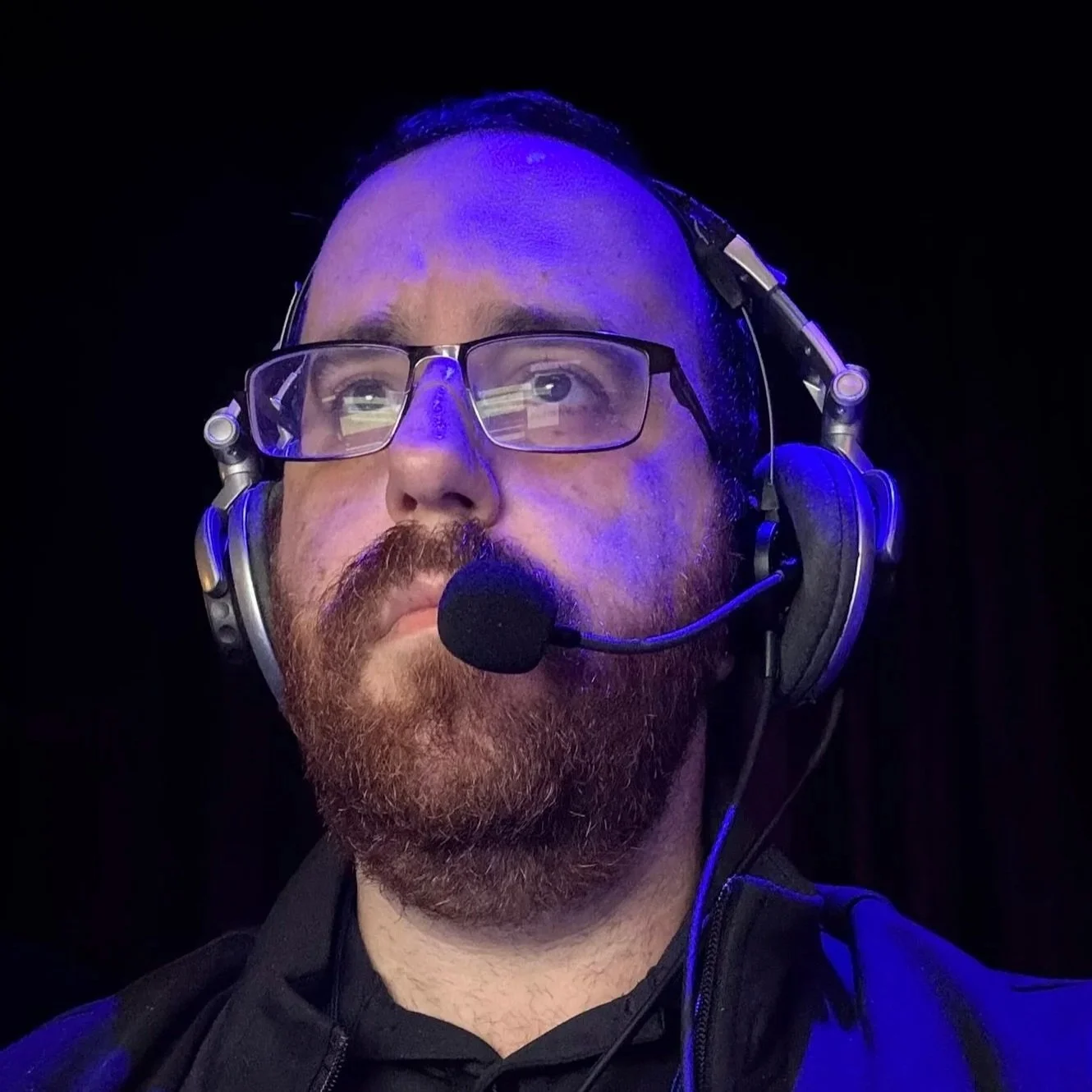 A man with glasses and a beard wearing a headset with microphone, looking up against a dark background.