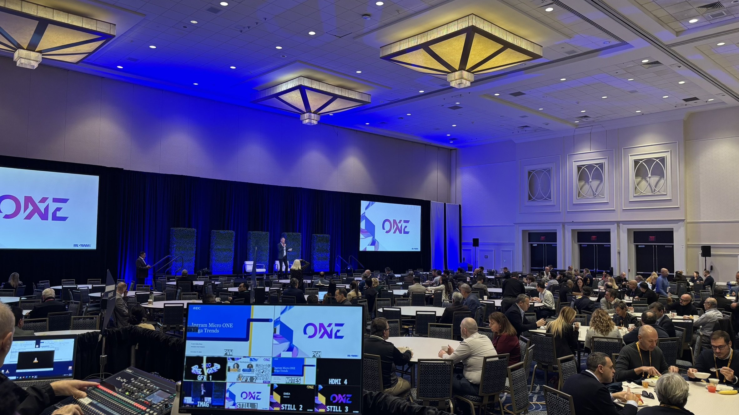 A large conference hall filled with round tables and people attending a professional event. There is a stage with a speaker, large screens displaying the word 'ONE,' and lighting with a blue hue. The room has high ceilings with decorative chandeliers.