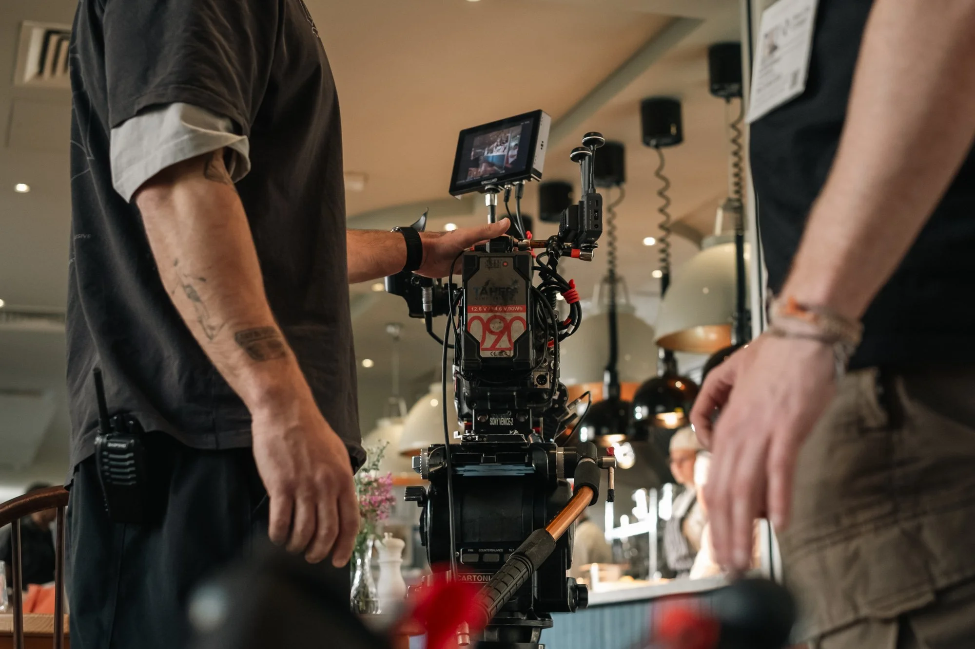 Two people operating a professional film camera inside a cafe, with one person pointing at a monitor attached to the camera.