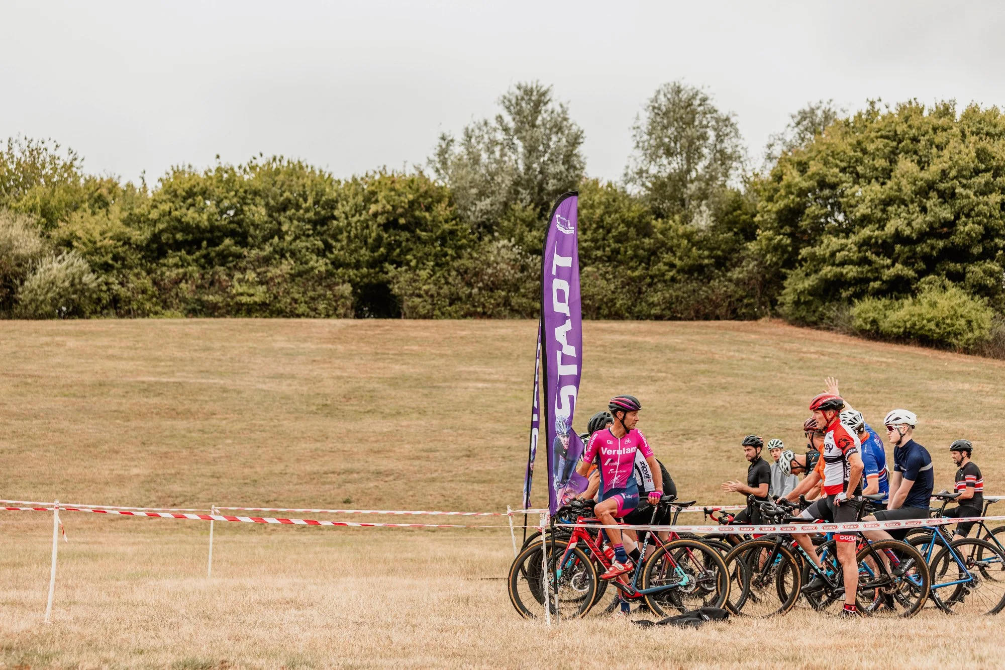 Group of cyclists gathered near a purple flag with a grassy hill and trees in the background during a cycling event.