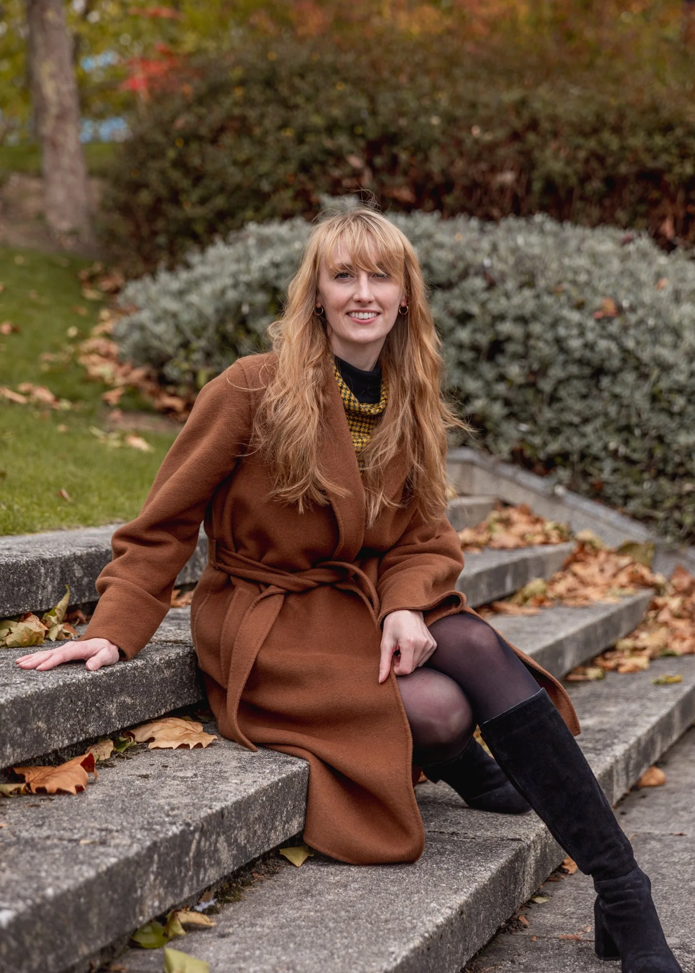 A woman with long wavy red hair, wearing a brown coat, black tights, and black knee-high boots, sitting on outdoor steps surrounded by autumn leaves and greenery.