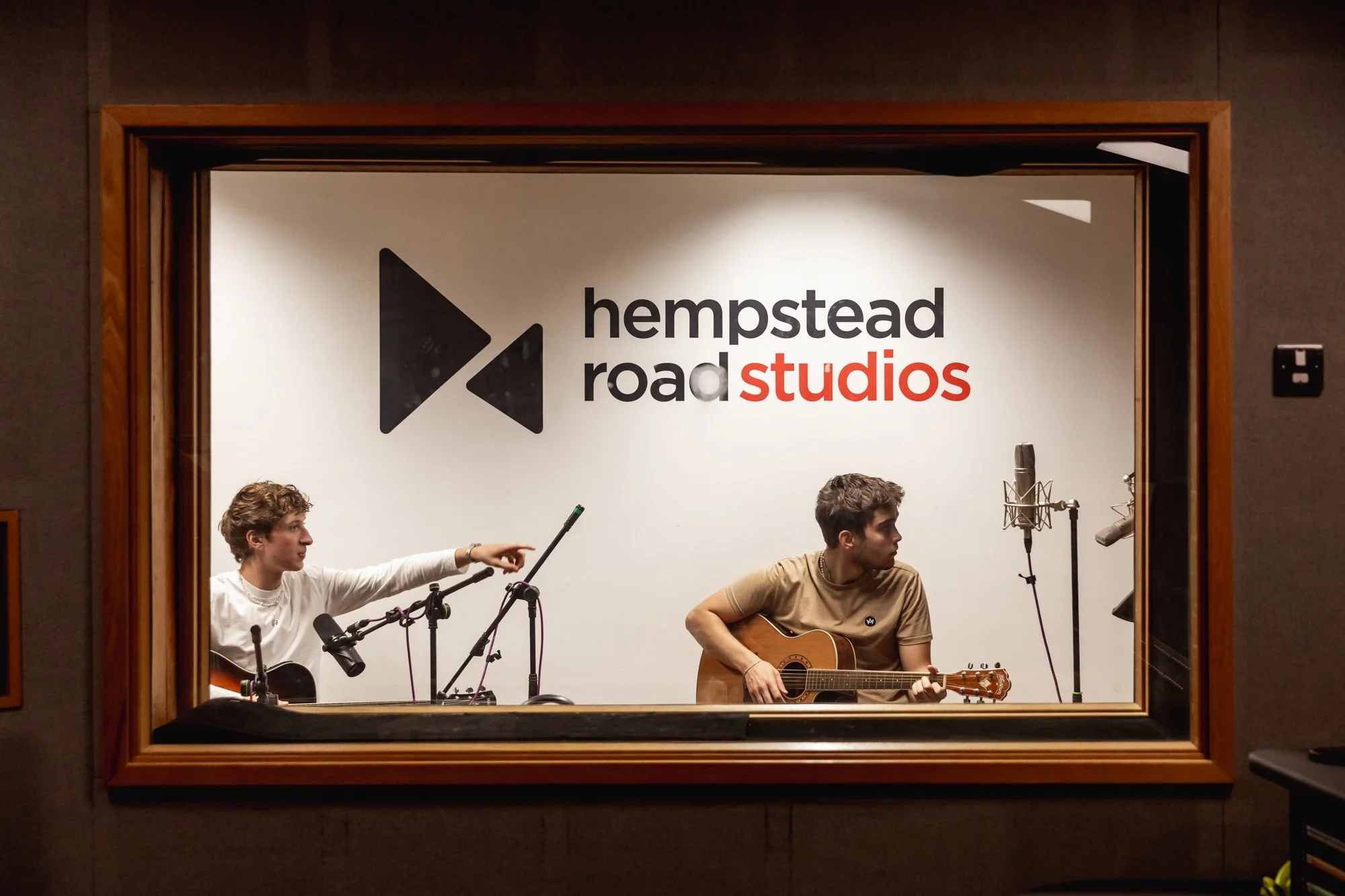 Two young men performing with guitars and microphones in a recording studio with a sign that reads "Hempstead Road Studios" in the background.